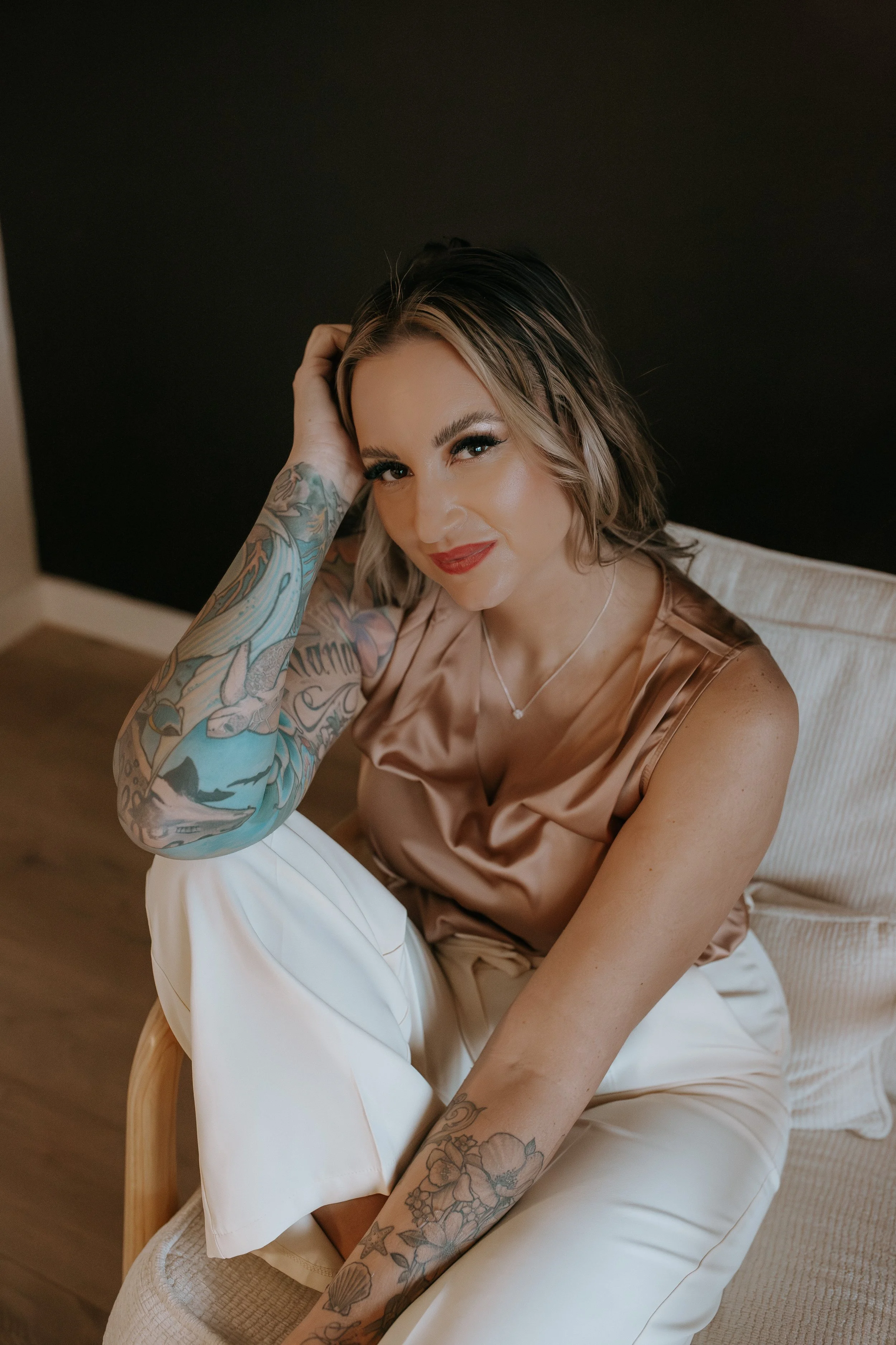 A woman with blonde hair and tattoos on her arms, wearing a silky beige top and white pants, sitting on a beige chair against a dark wall, smiling and looking at the camera.