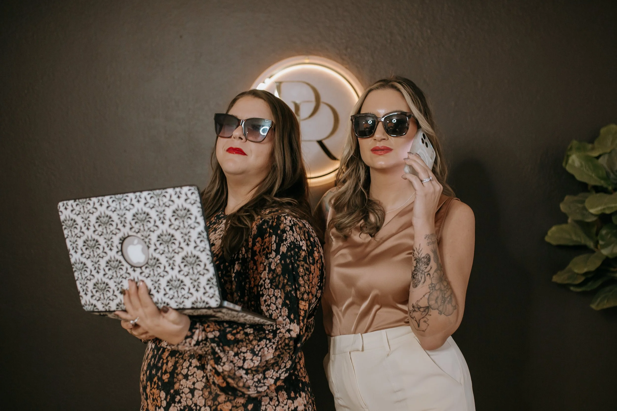 Two women with sunglasses, one holding a laptop and the other talking on a cell phone, standing in front of a dark wall with backlit logo.