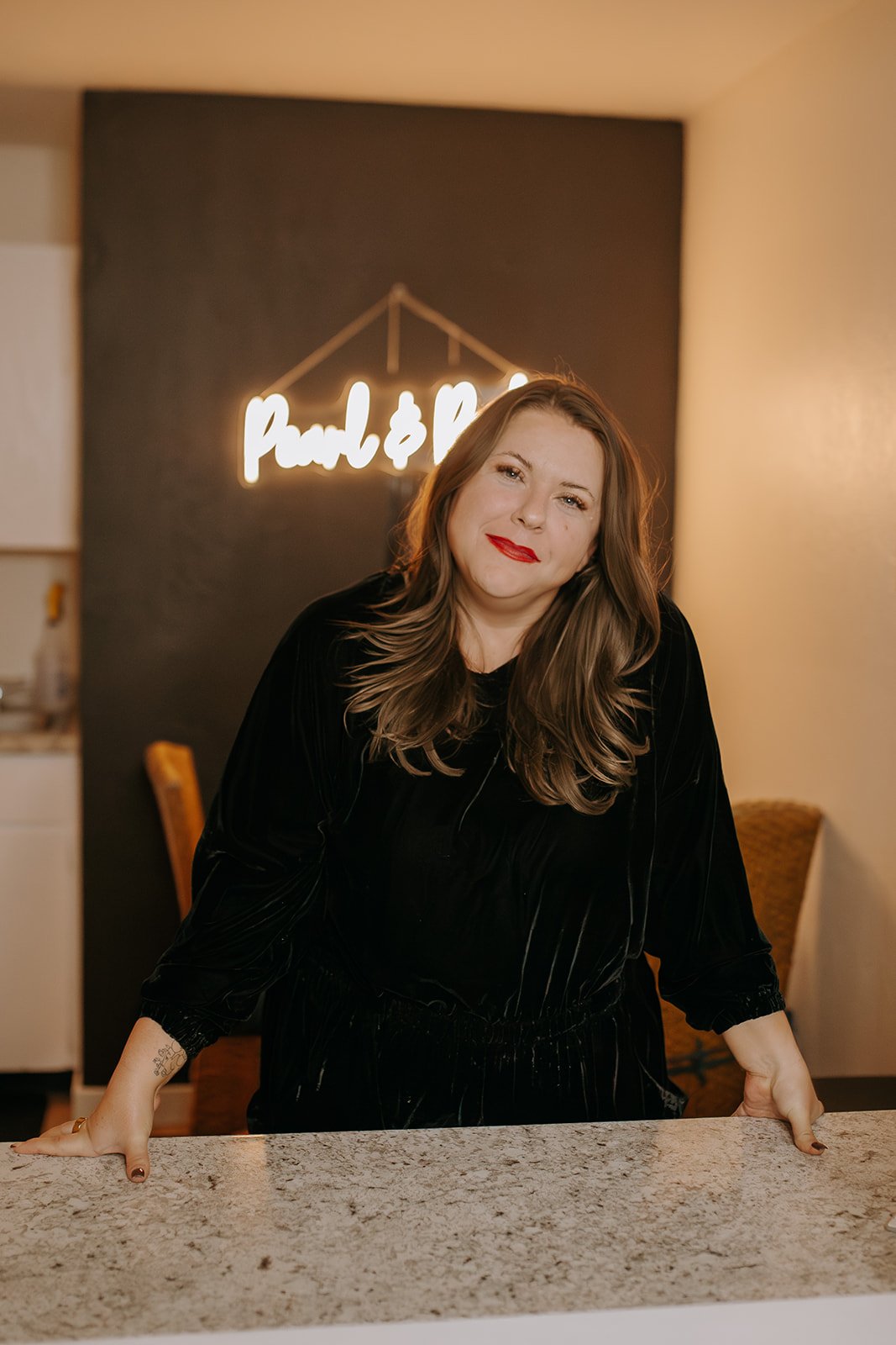 A woman with wavy brown hair and red lipstick leaning on a kitchen counter in front of a dark wall with a neon sign that says 'punch & pie'.
