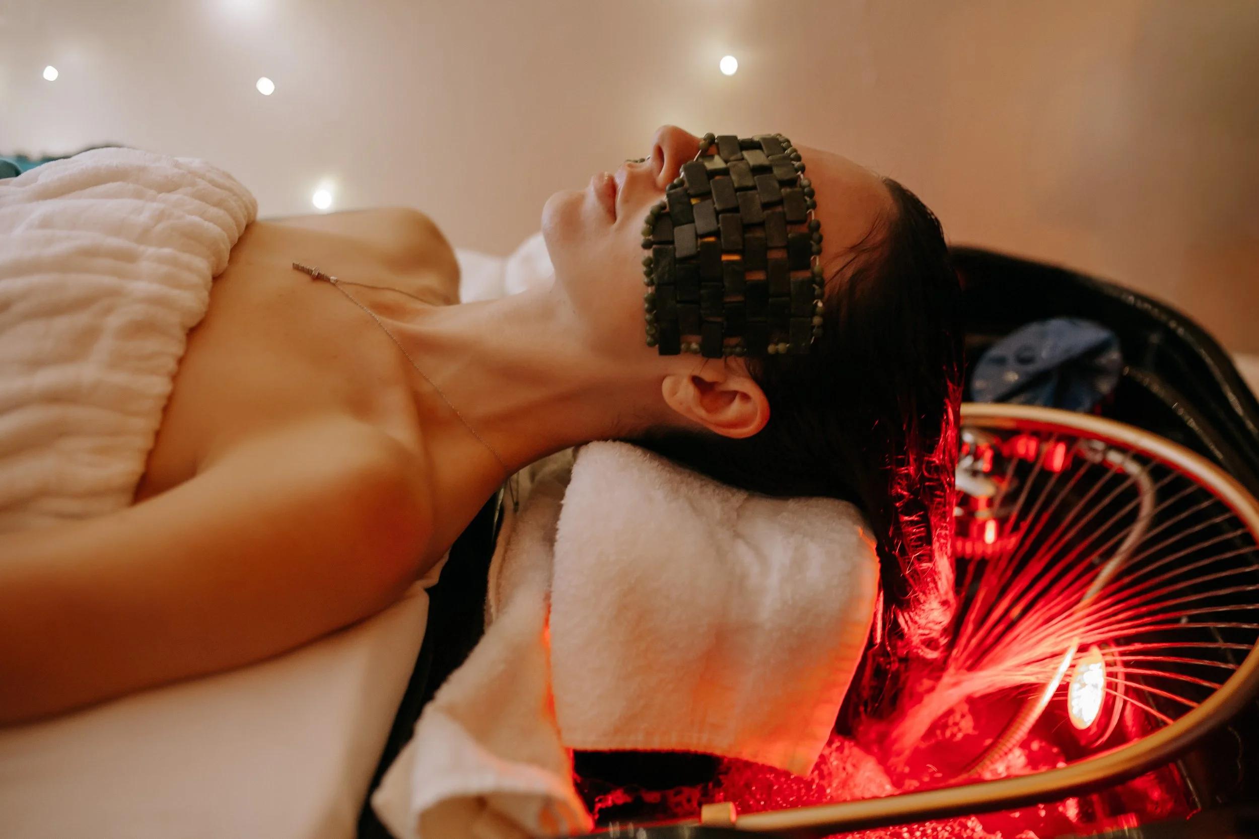 A person lying on a bed with a massage or spa treatment, wearing a thermal or heat therapy device on their face, covered with a beige towel, with red light and a fan in the background.