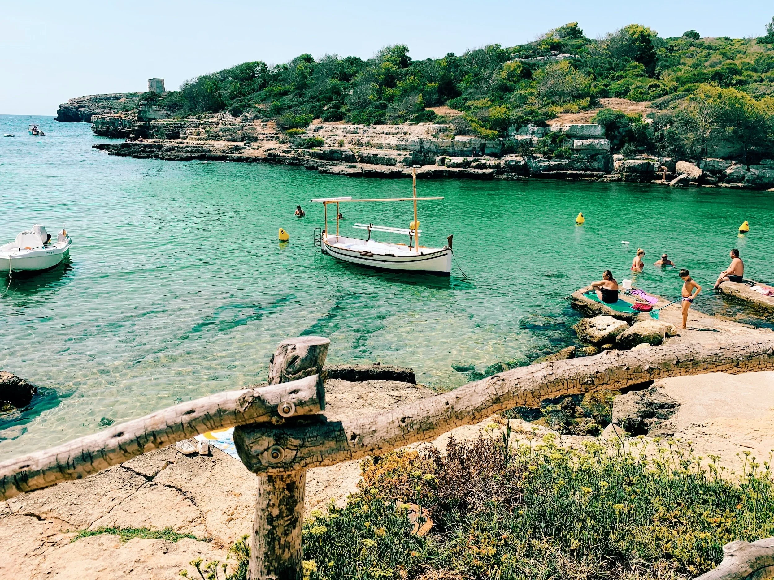 People swimming and relaxing by a rocky shoreline in a clear, greenish blue bay, with small boats anchored nearby and a lush, green hillside in the background.