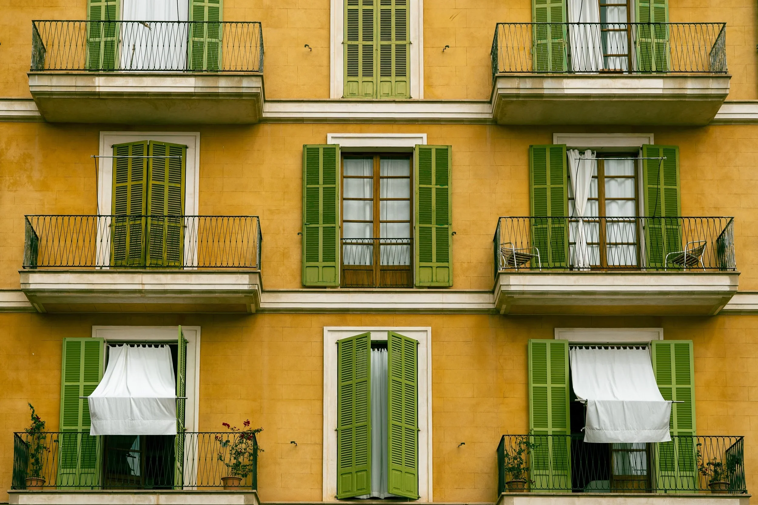 Yellow building with green French shutters, some open, some closed, and white awnings over balconies decorated with plants.