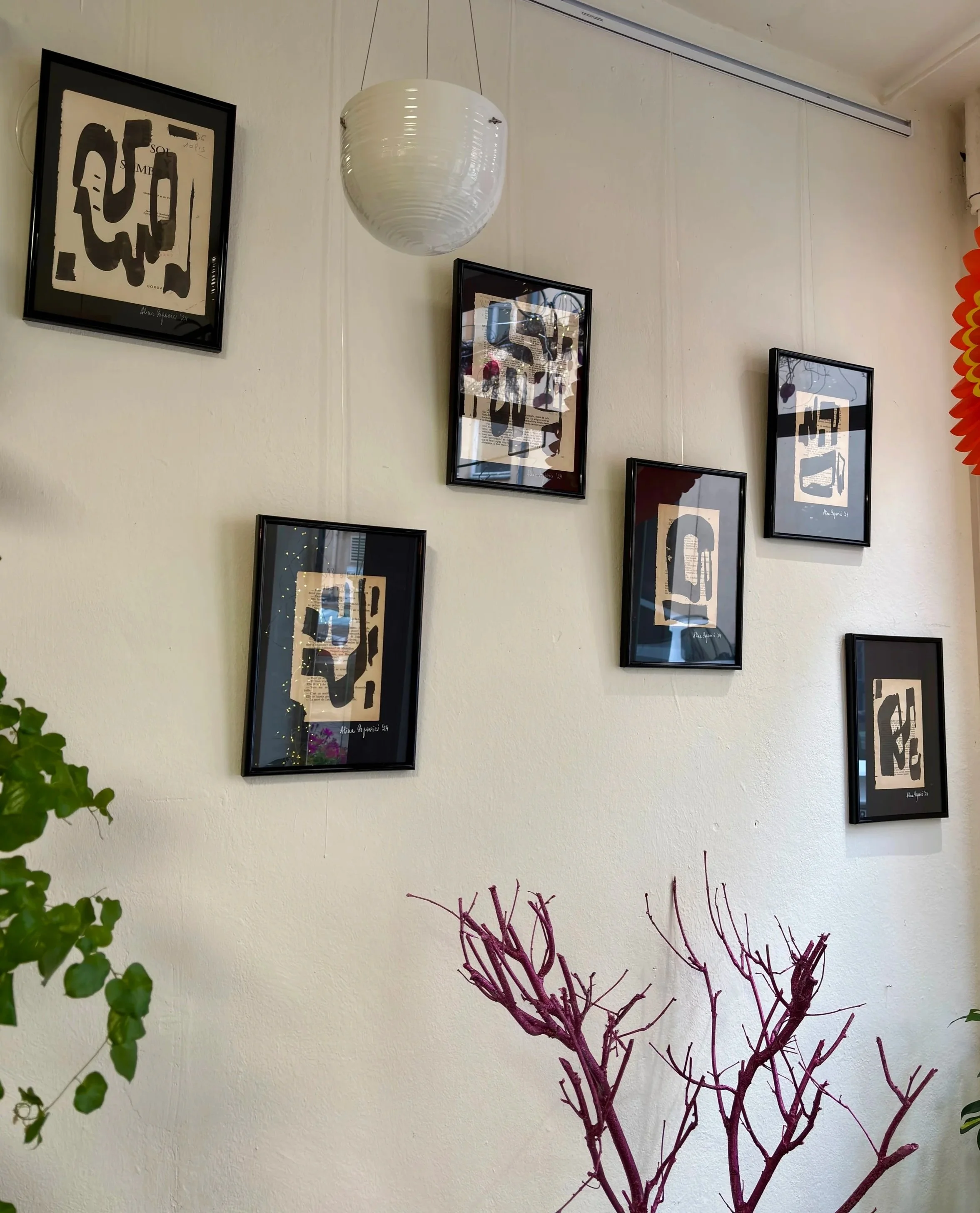 Decorative wall with six framed abstract art prints arranged in two rows, a white hanging paper lantern, and a few plants including a leafless branch painted purple in the foreground.