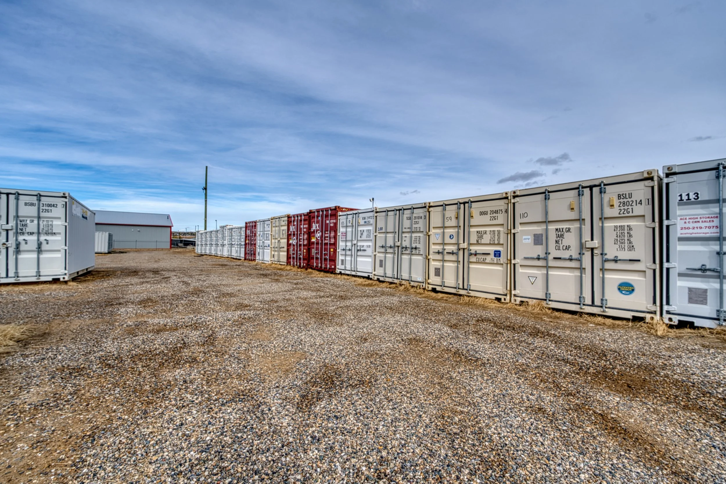 Row of white and red shipping containers on a gravel lot under a cloudy sky.