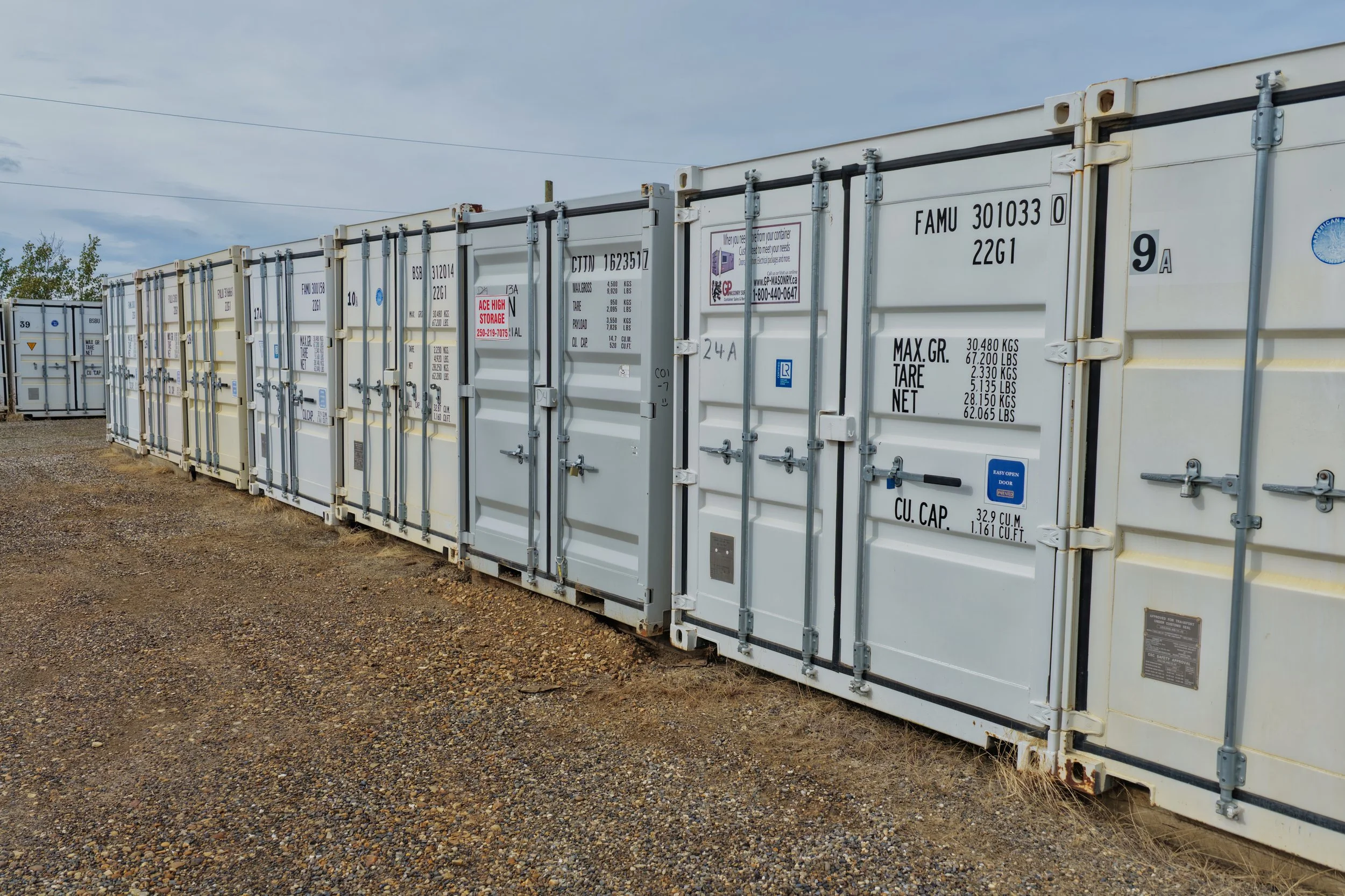 Row of white storage containers lined up outdoors on a gravel surface under a cloudy sky.