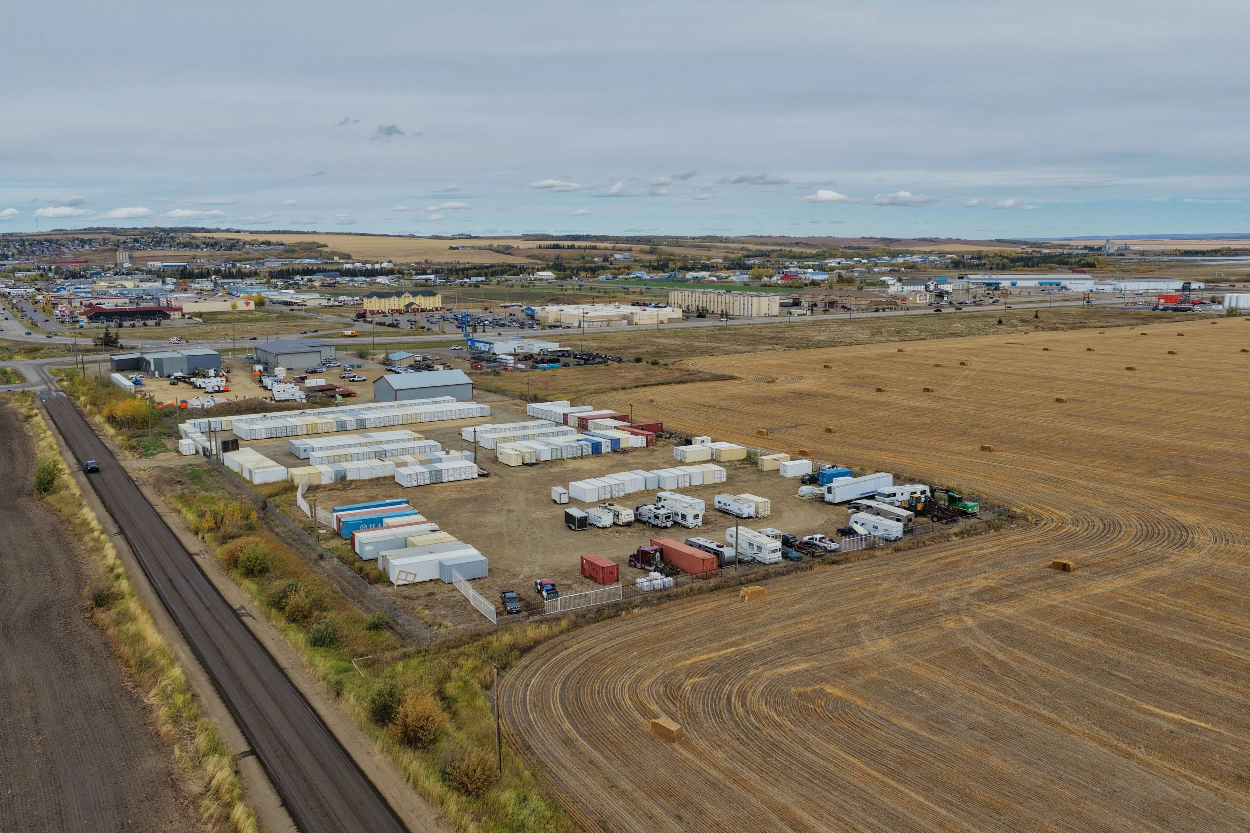 Aerial view of a populated area with a large parking lot containing delivery trucks and storage containers next to agricultural fields and a cityscape in the background.