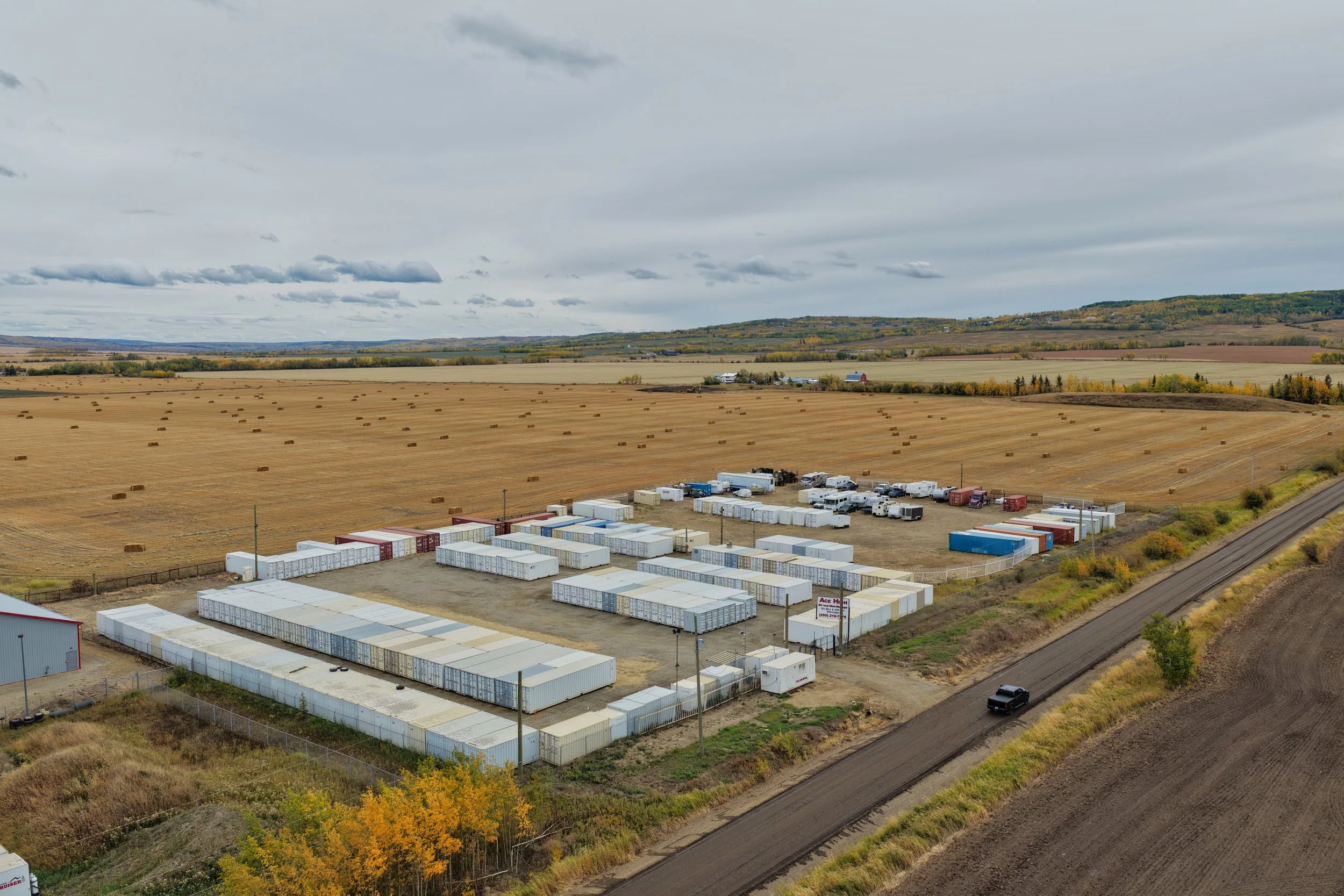 Aerial view of a storage facility with numerous shipping containers and trucks, surrounded by farmland with hay bales and rolling hills in the background during daytime.