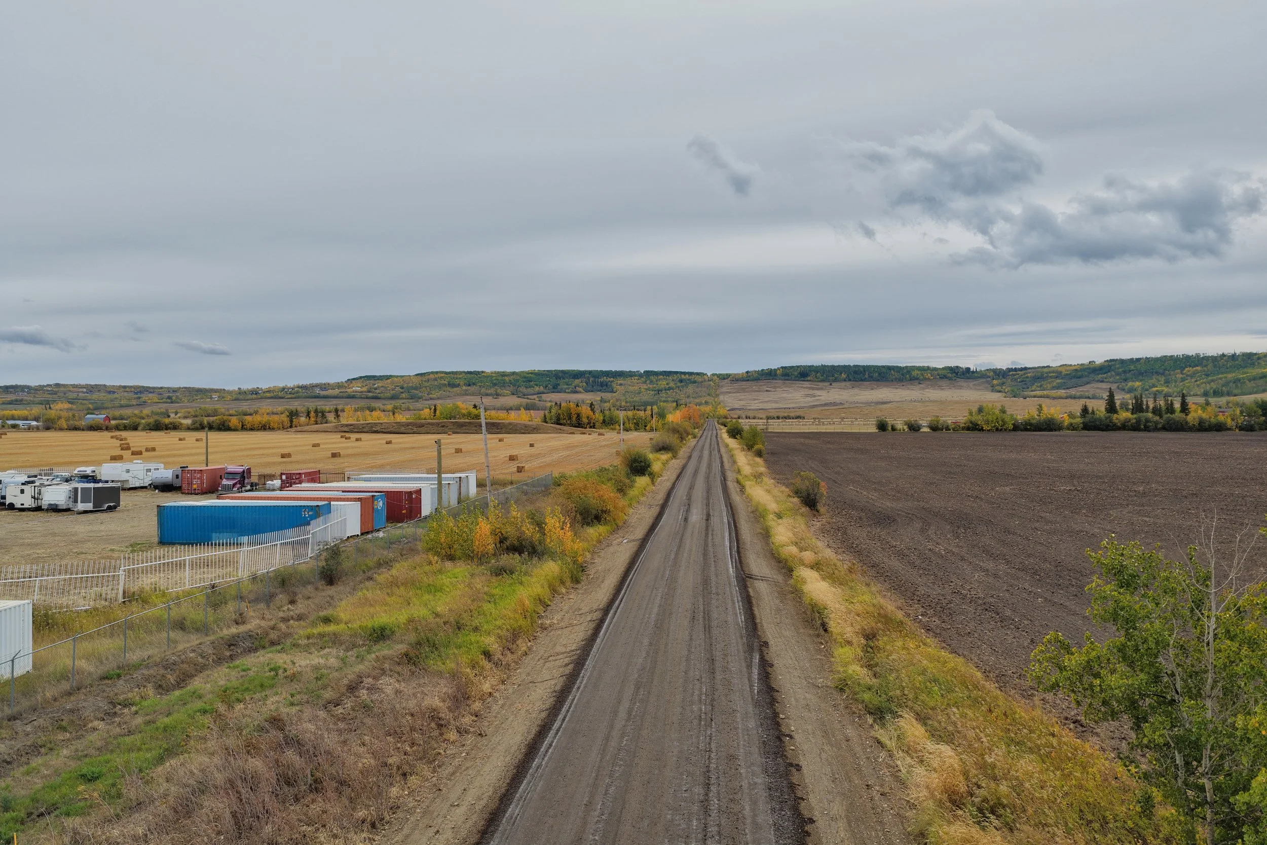 Long dirt road stretching into the horizon with agricultural fields and containers on the left side, overcast sky above.