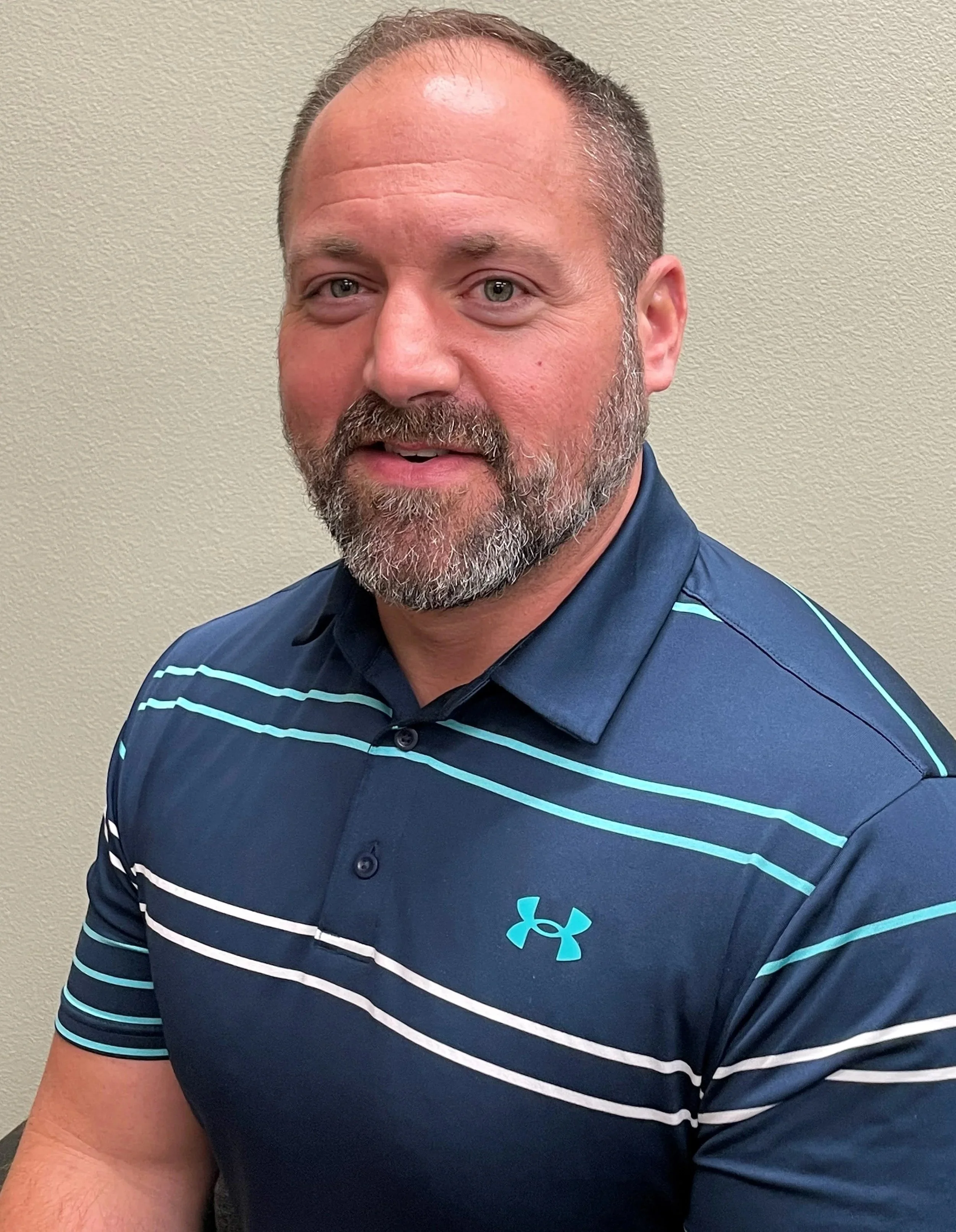 A middle-aged man with a beard and short hair, wearing a navy blue Under Armour polo shirt with light blue and white stripes, posing in front of a beige wall.
