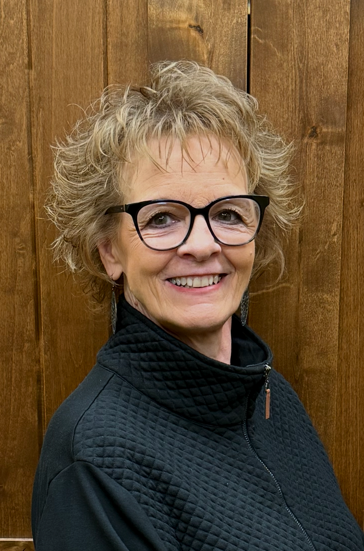 A woman with curly short hair wearing glasses and earrings, smiling, dressed in black top, standing indoors with a plant and blurred background.
