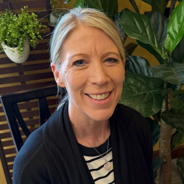 A smiling woman with blonde hair and blue eyes, wearing a black and white striped shirt and a black cardigan, seated indoors near green houseplants.