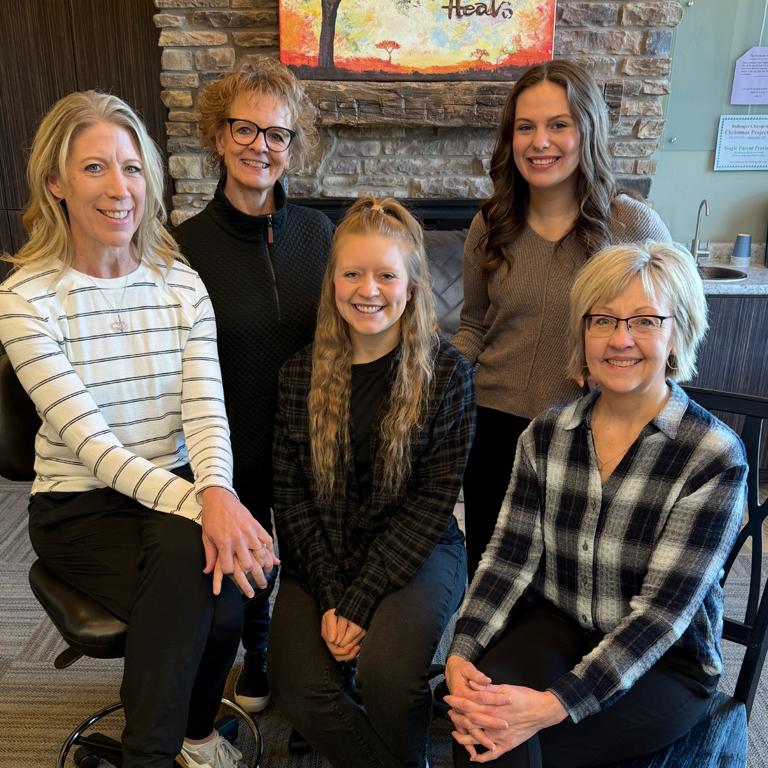 Five women smiling at the camera in a cozy room with a stone fireplace and artwork behind them.