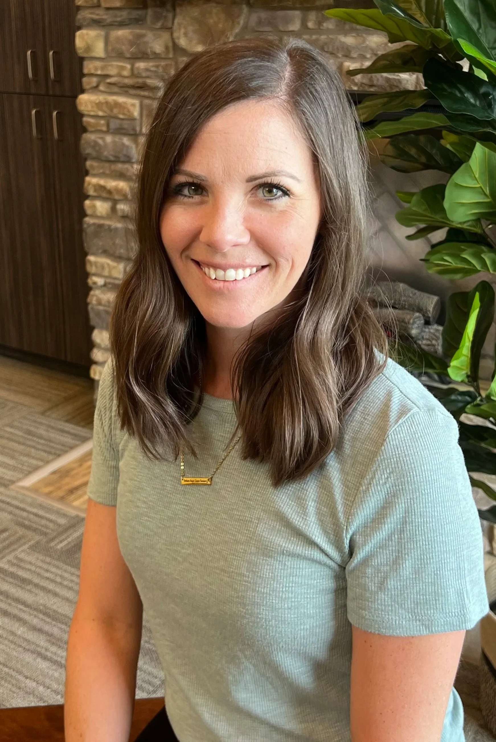A woman with shoulder-length brown hair, smiling, wearing a light gray t-shirt and a gold necklace, indoors with a stone wall and green plant in the background.