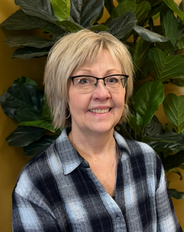 A smiling woman with short blonde hair and glasses, wearing a rust-colored sweater and a long gold necklace, sitting near green plants.