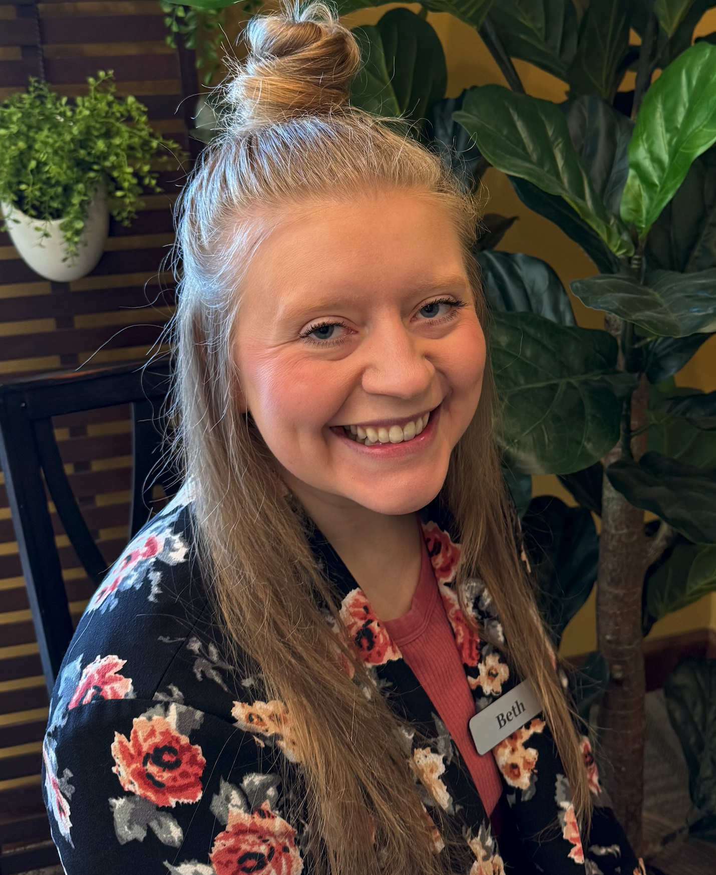 A smiling woman with long blonde hair tied up in a top bun, wearing a floral blazer and a pink top, sitting in front of indoor plants.