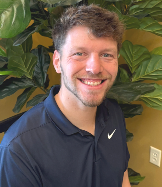 A smiling man with short brown hair, a beard, and blue eyes, wearing a navy blue Nike polo shirt, stands in front of a large leafy green plant and a yellow wall.