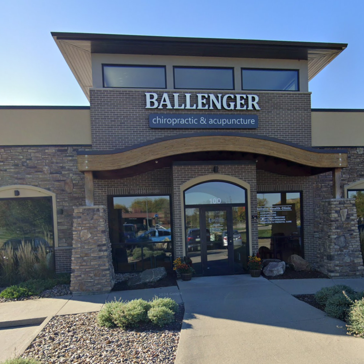 Exterior of a chiropractic and acupuncture clinic named Ballenger with signs and a glass door entrance, surrounded by landscaped plants and rocks.