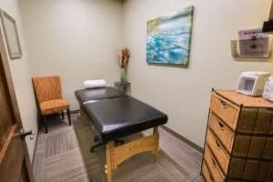 A massage room with a massage table, a wooden dresser, a chair, and beach artwork on the wall