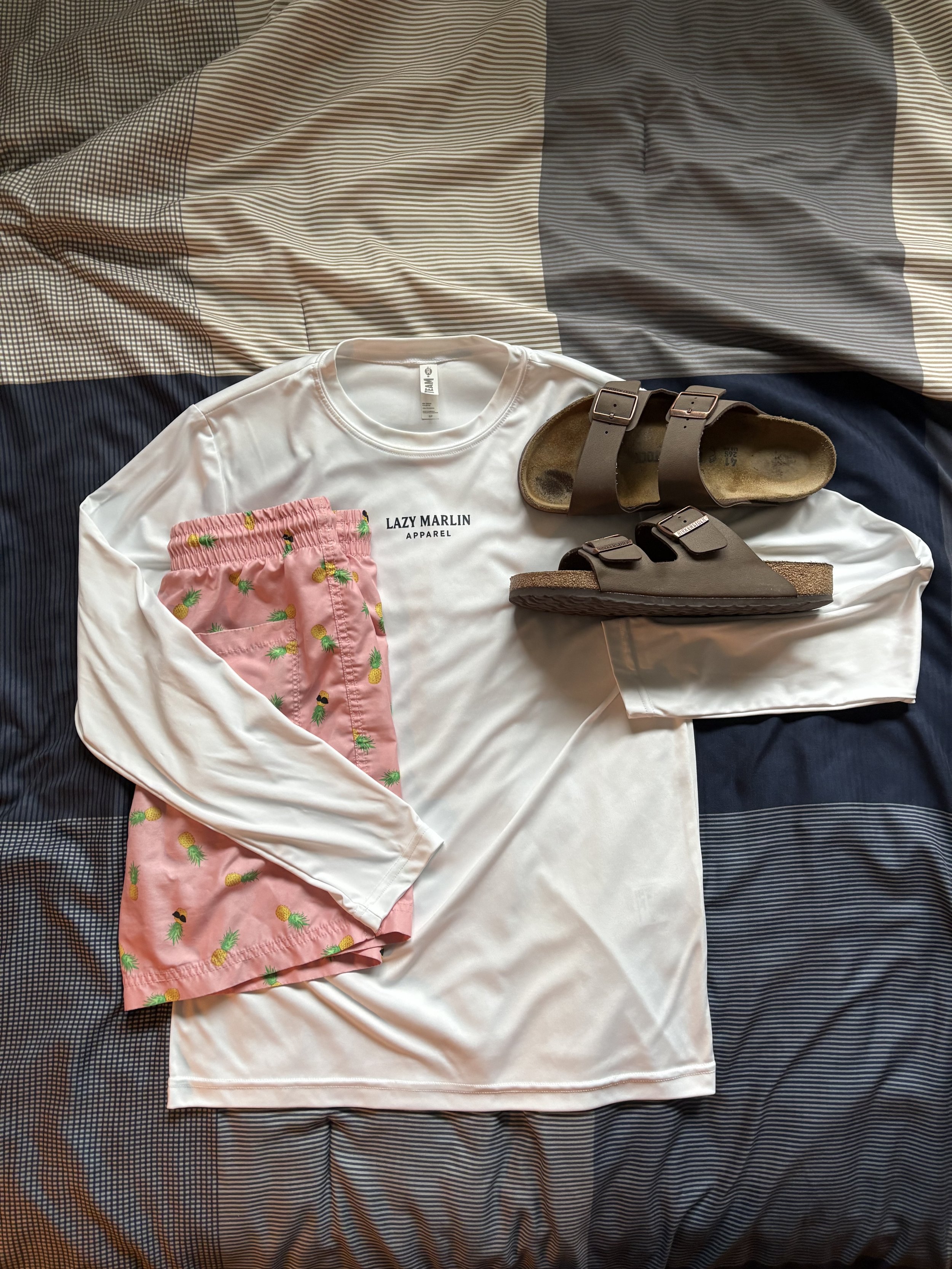 White long-sleeve shirt with 'Lazy Marlin Apparel' text, pink shorts with pineapple print, and two pairs of brown sandals on a bed with striped bedding.