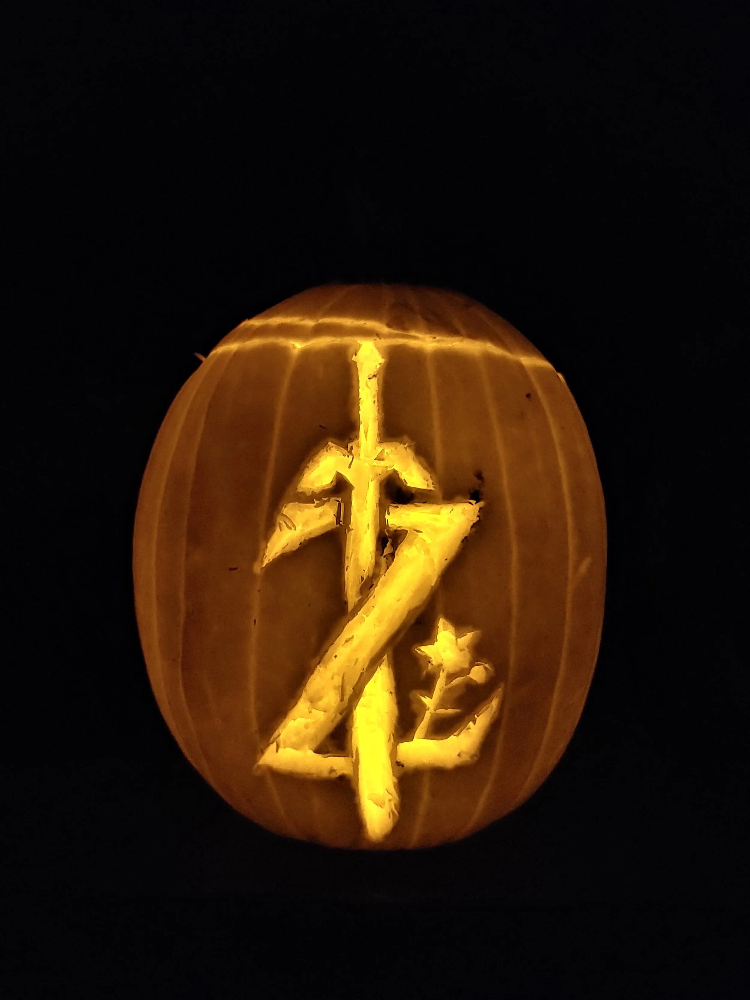 A carved pumpkin with a design of the Legend of Zelda "Z" with Master Sword through the top, illuminated from inside against a black background.
