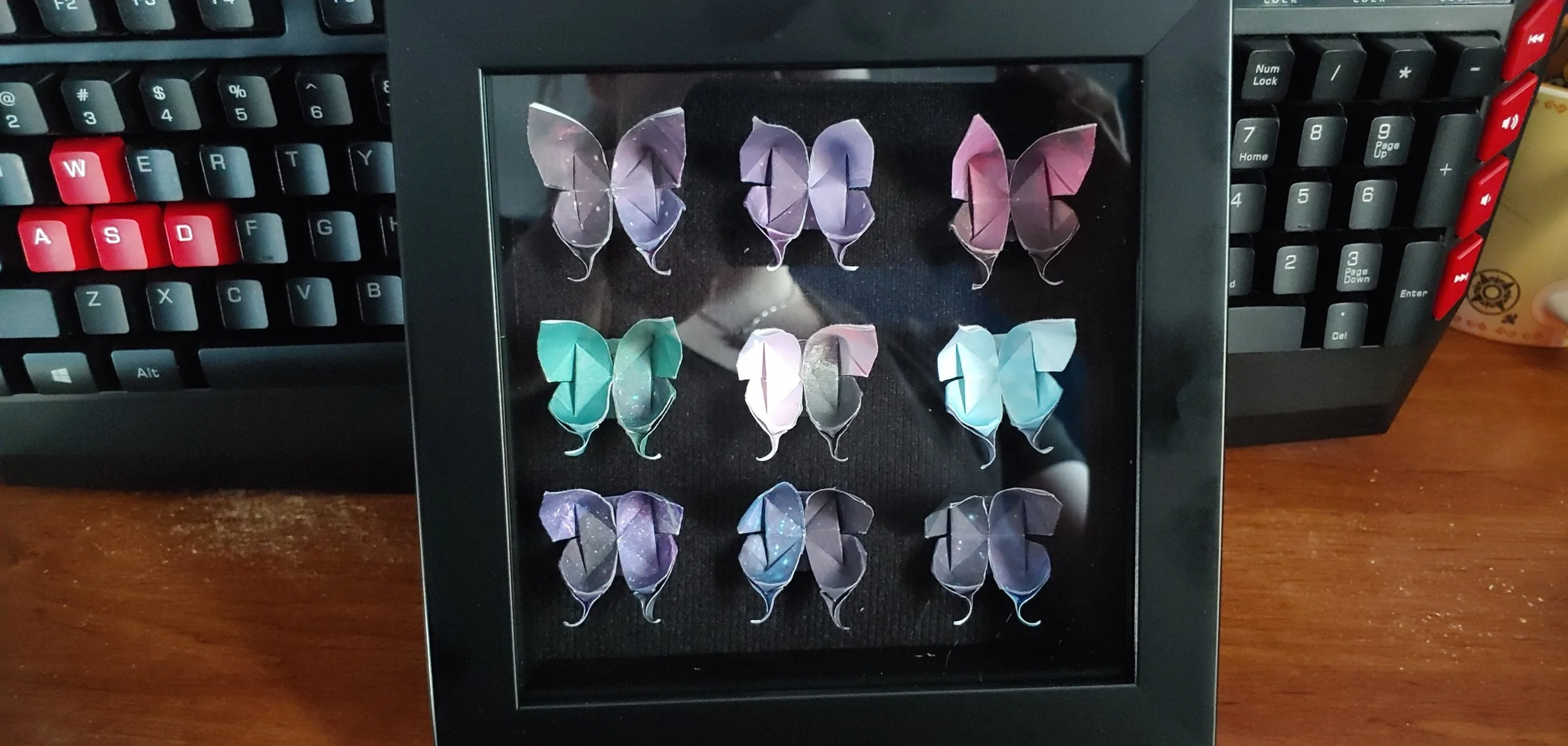 A shadow box frame with 9 colourful origami butterflies arranged in three rows, placed on a dark surface. The frame is on a wooden desk, with a black keyboard with red keycaps visible in the background.