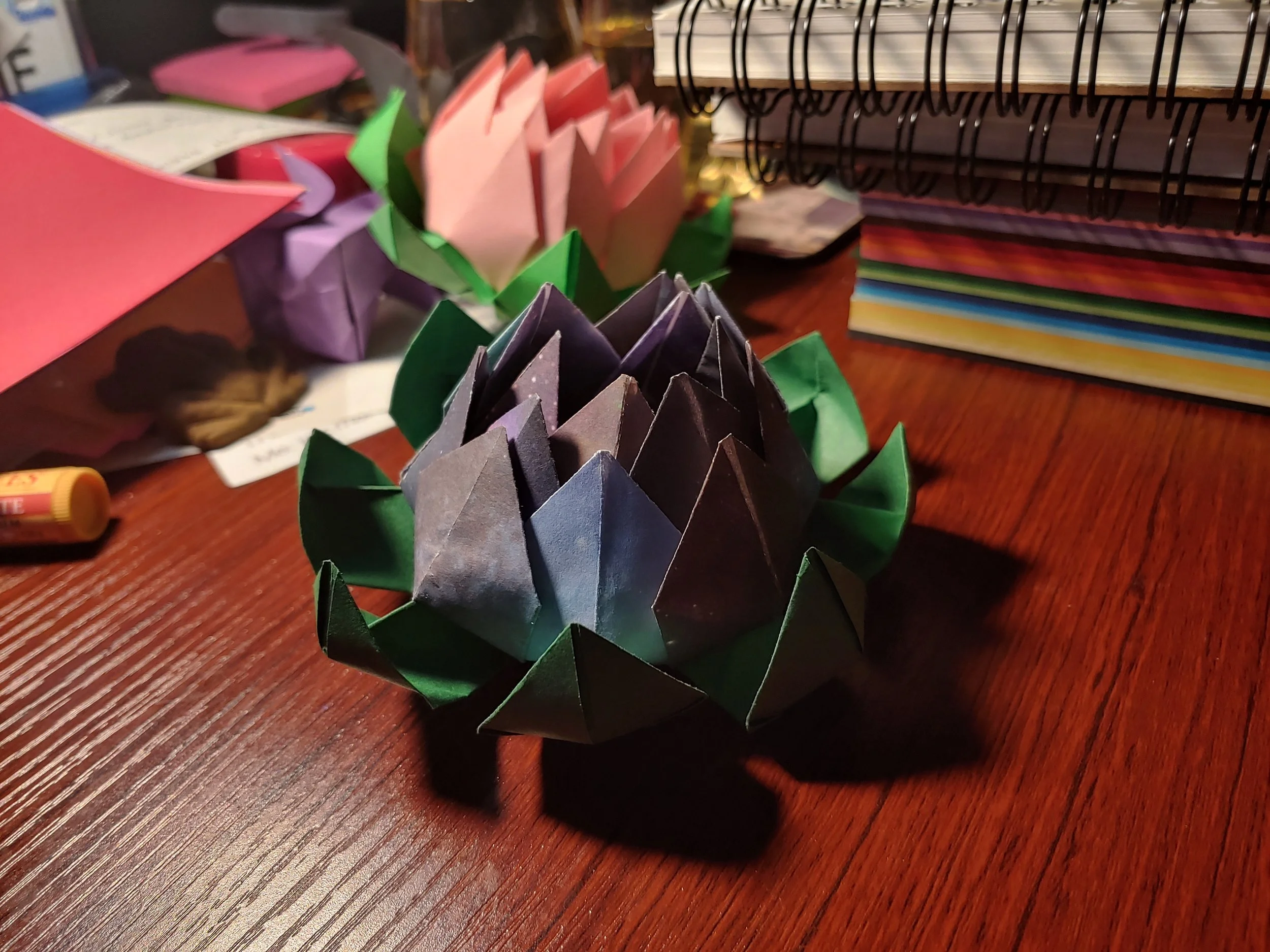 Colourful origami lotuses with green leaves on a wooden desk, stacks of notebooks and spiral-bound books in the background.
