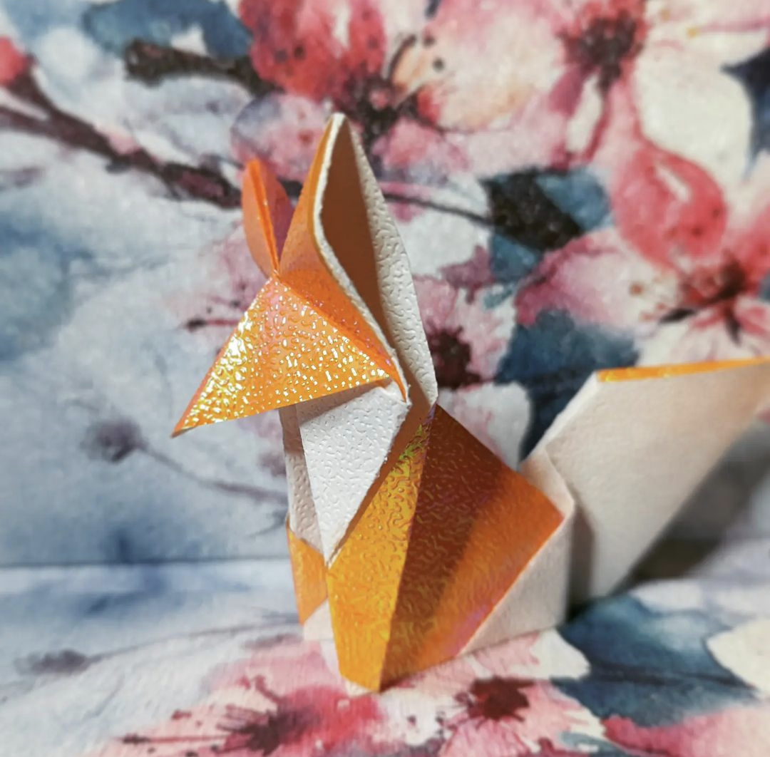 Close-up of an origami fox made from textured orange and white paper, set against a floral watercolour background.