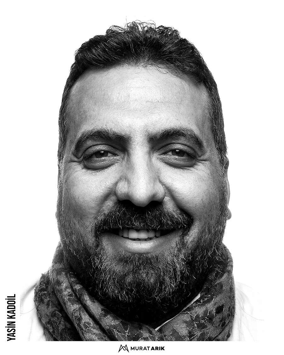 Close-up black-and-white portrait of a smiling man with a beard and mustache, wearing a scarf.