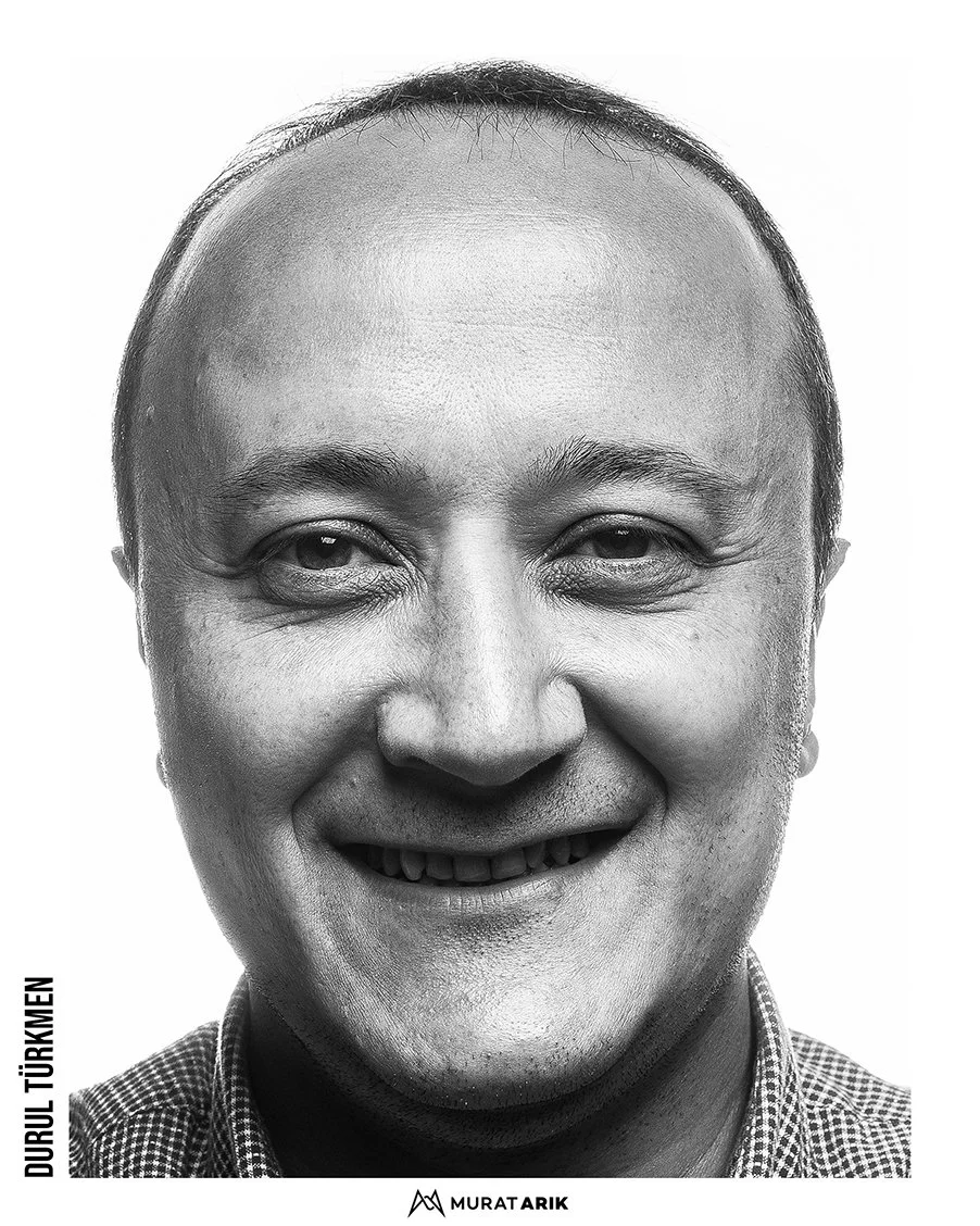 Close-up black and white portrait of a smiling man with a bald head, wearing a checkered shirt.