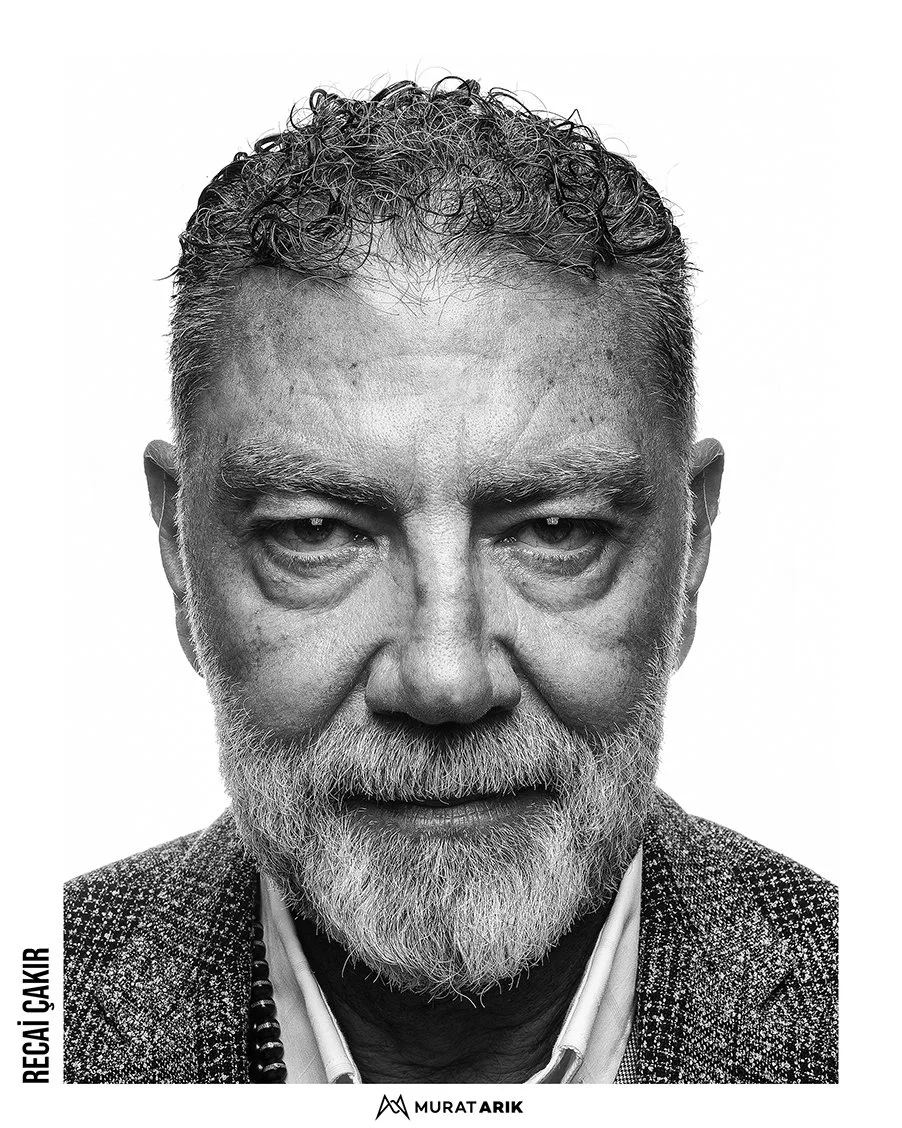 Close-up of an older man with curly hair and a beard, dressed in a textured blazer and shirt, looking directly at the camera.