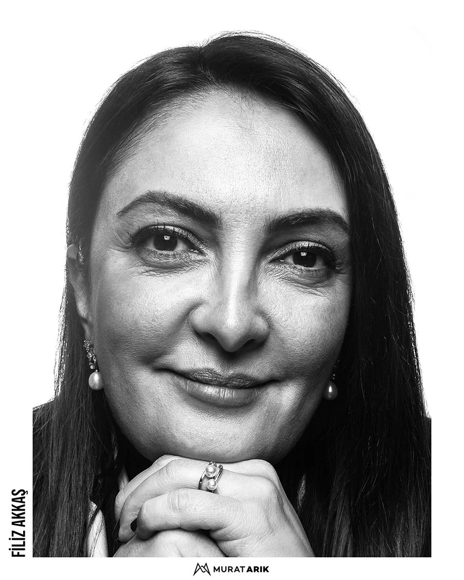 Black and white close-up portrait of a woman with long dark hair, wearing earrings and a ring, smiling with hands clasped under her chin.