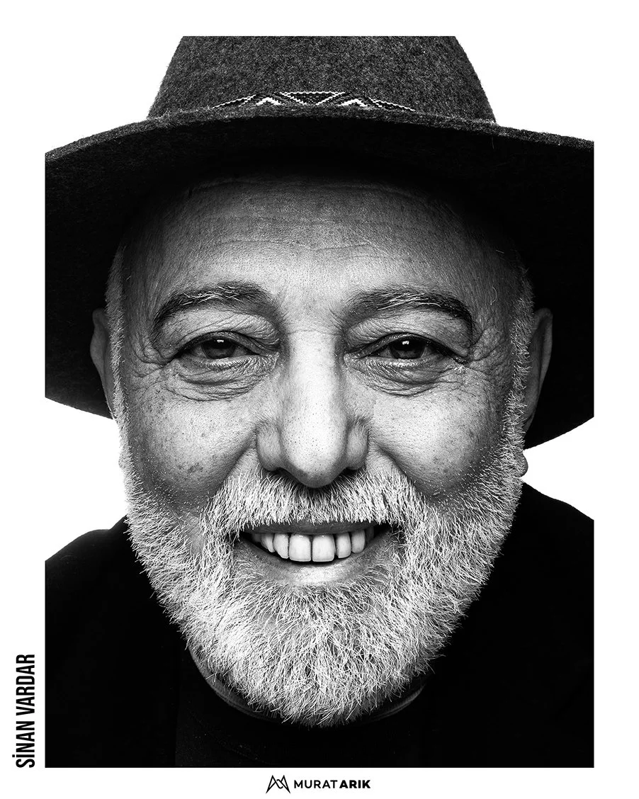 Close-up black and white portrait of Sinan Wardar, an older man with a beard, smiling, wearing a dark hat, with his face filling the frame.