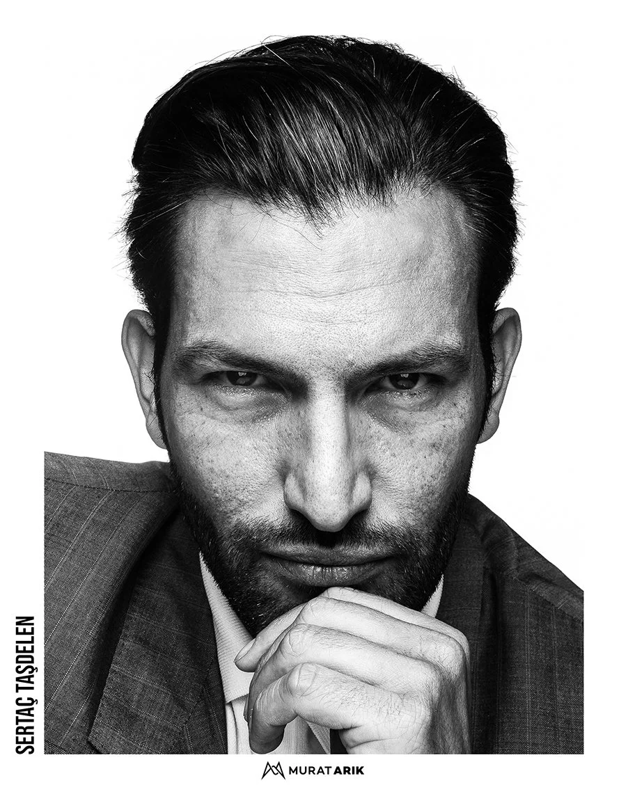 Close-up of a man with dark hair and a beard, wearing a suit, looking serious and holding his chin with his hand. Black and white photo.