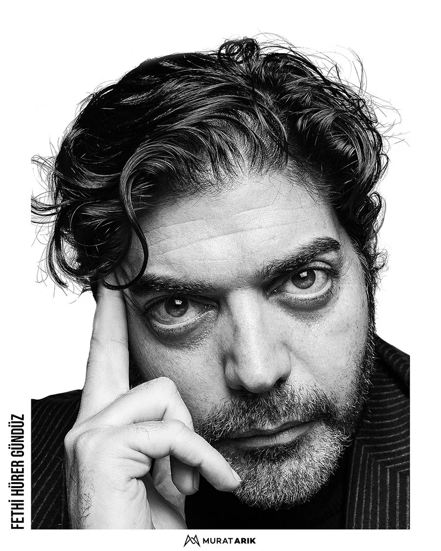 Black and white close-up photograph of a man with wavy hair and a beard, looking directly at the camera with his hand resting on his temple.