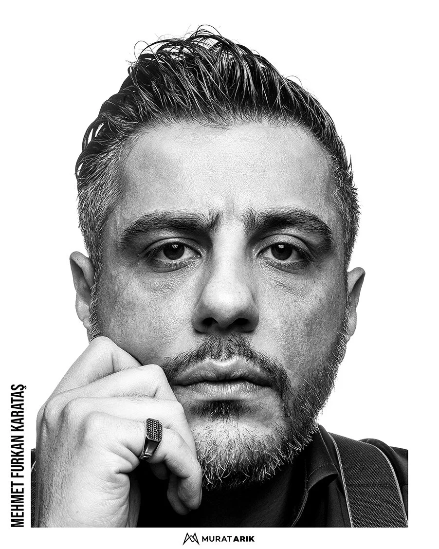 Black and white close-up portrait of a man with styled hair and a beard, resting his chin on his hand, wearing a ring and a dark shirt, with a serious expression.