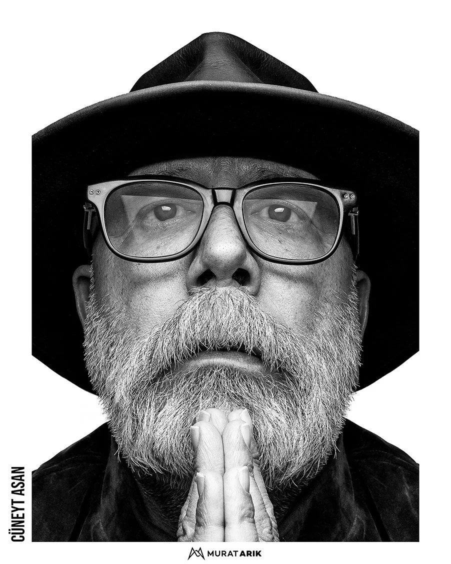 Close-up black and white portrait of a bearded man wearing glasses and a wide-brimmed hat, with hands pressed together in prayer or meditation. The man has a serious expression.