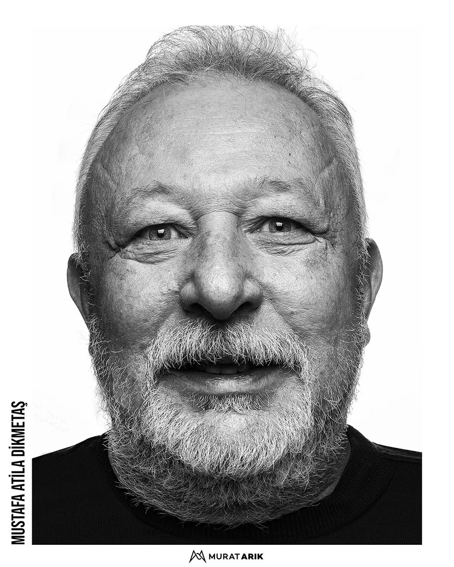 Black and white portrait of Mustafa Atila Dikmetas smiling, with gray hair and beard, wearing a black shirt.
