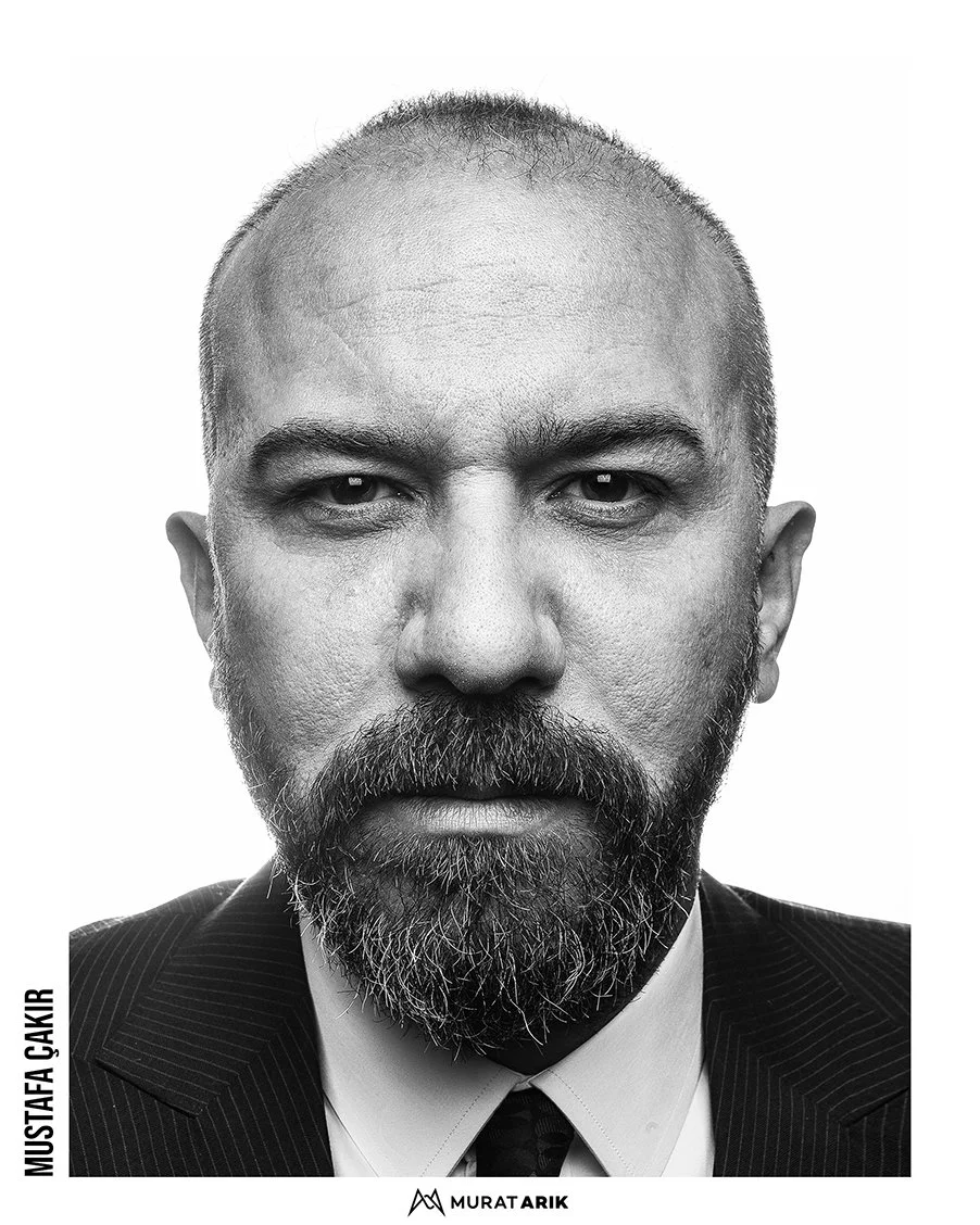 Black and white close-up portrait of a man with a beard and short hair, wearing a suit and tie, looking directly at the camera.