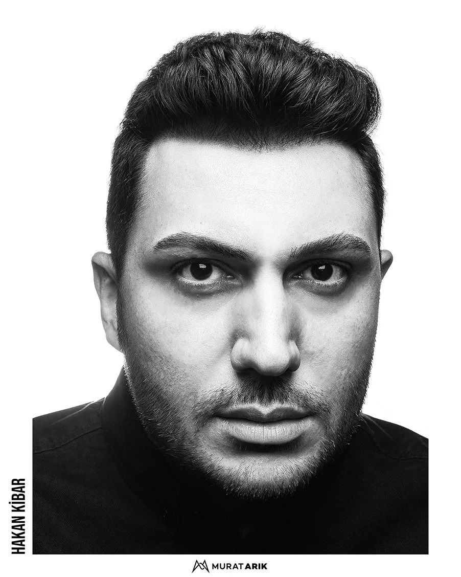 Black and white close-up portrait of a man with dark hair styled upward, looking directly at the camera with intense eyes, wearing a dark shirt.