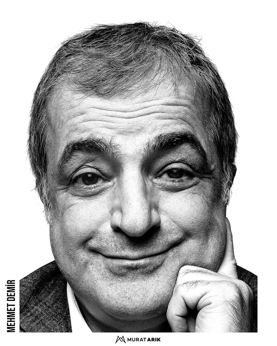 A black and white portrait of a smiling middle-aged man with gray hair, resting his chin on his hand, wearing a blazer.