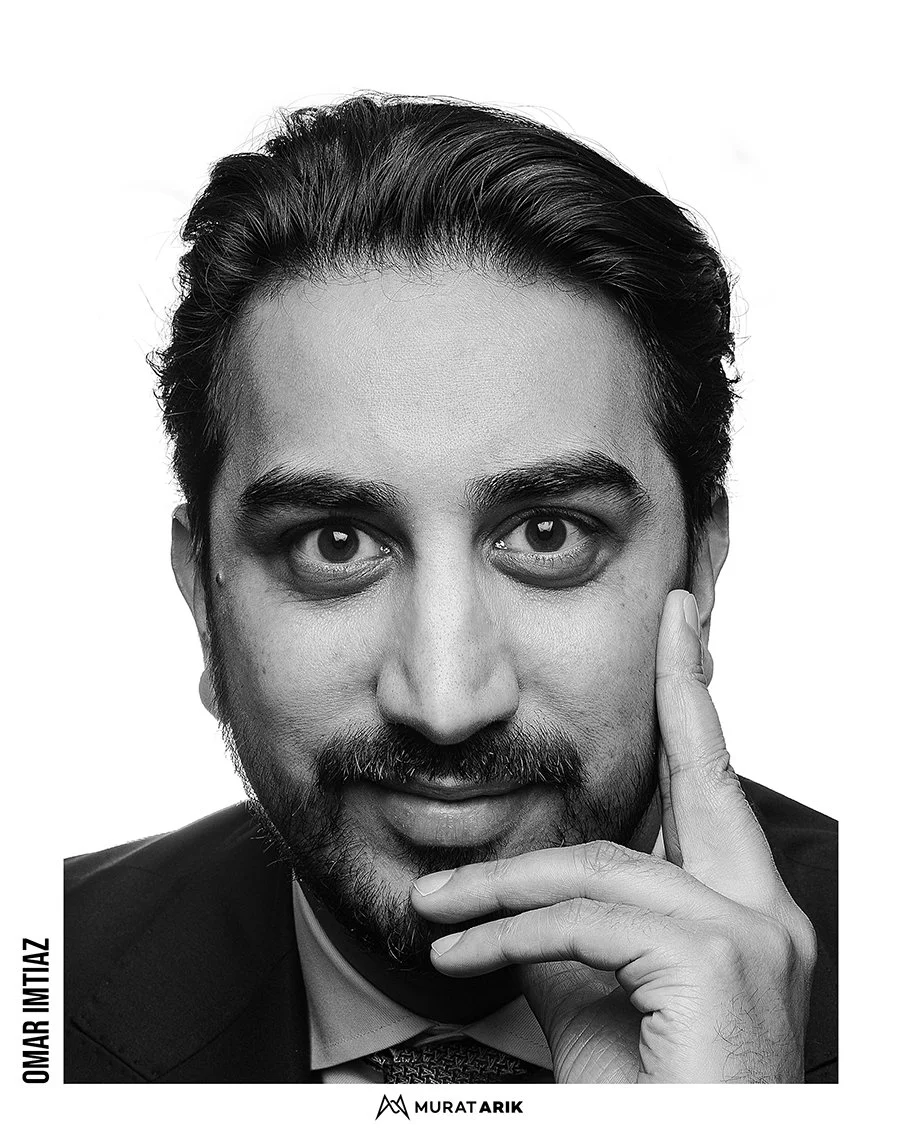 A black and white portrait of Omer Imtiaz, a man with dark hair, beard, and mustache, wearing a suit, with his hand resting on his face.