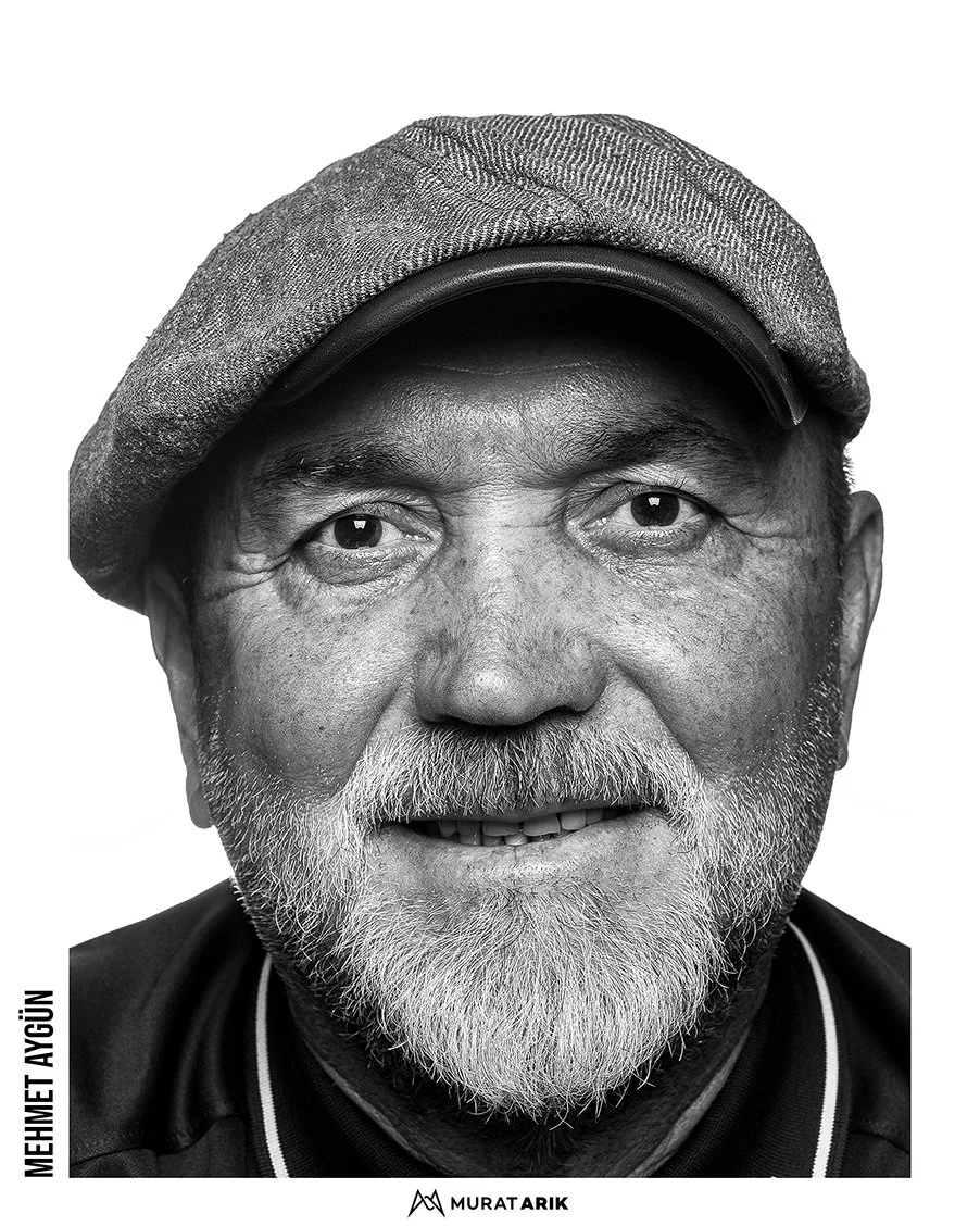 Close-up black and white portrait of a smiling older man with a beard, wearing a flat cap and a dark jacket.
