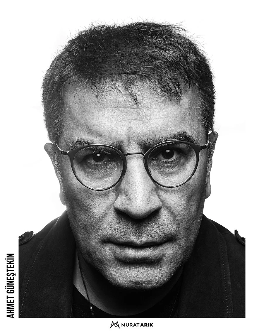 Black and white close-up portrait of Ahmet Güneytekin wearing round glasses, with a serious expression, short dark hair, and a dark shirt.