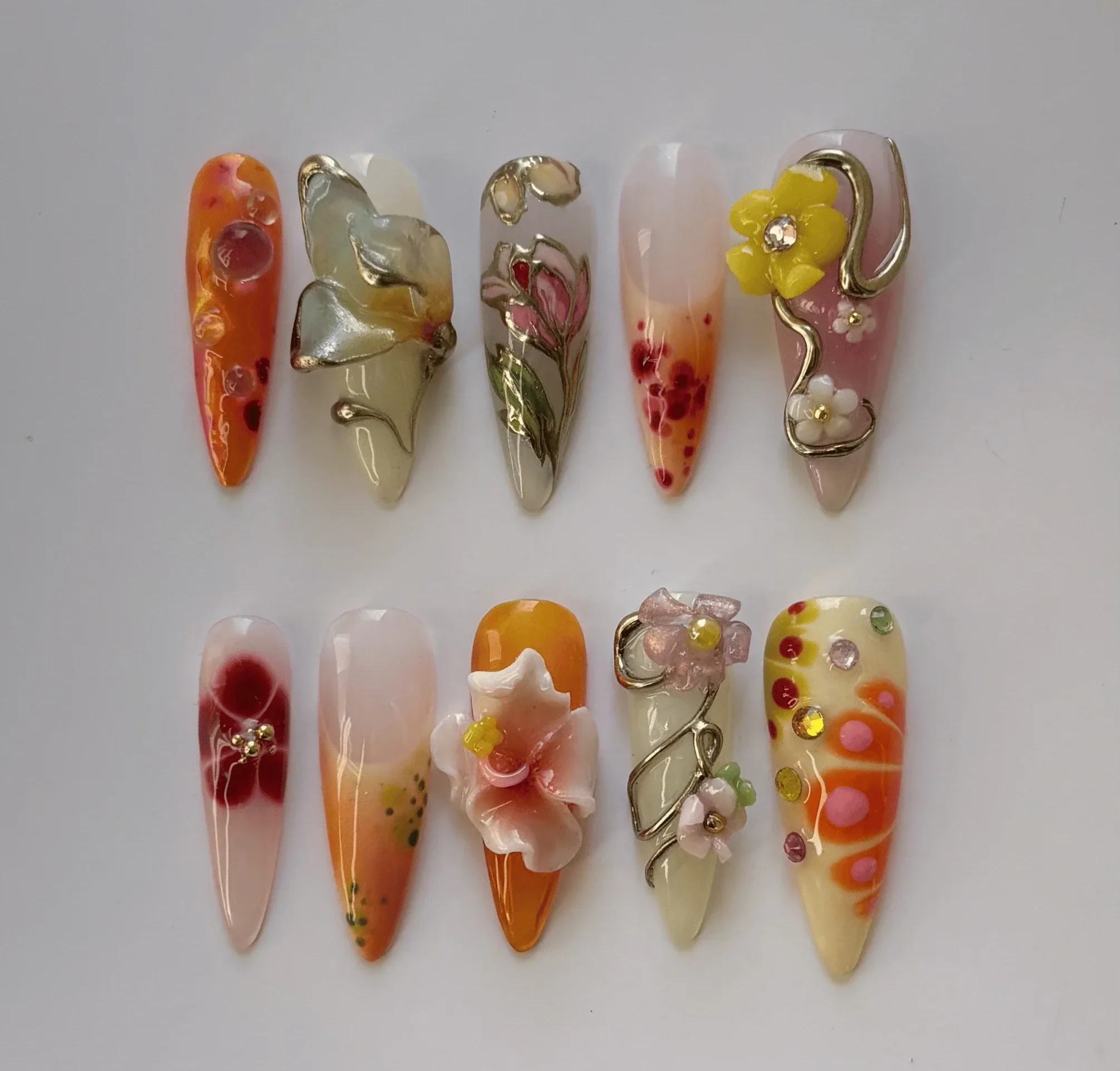 Dix ongles artificiels décorés avec des motifs floraux et colorés, comportant des embellissements en métal, perles et strass, disposés sur un fond blanc.