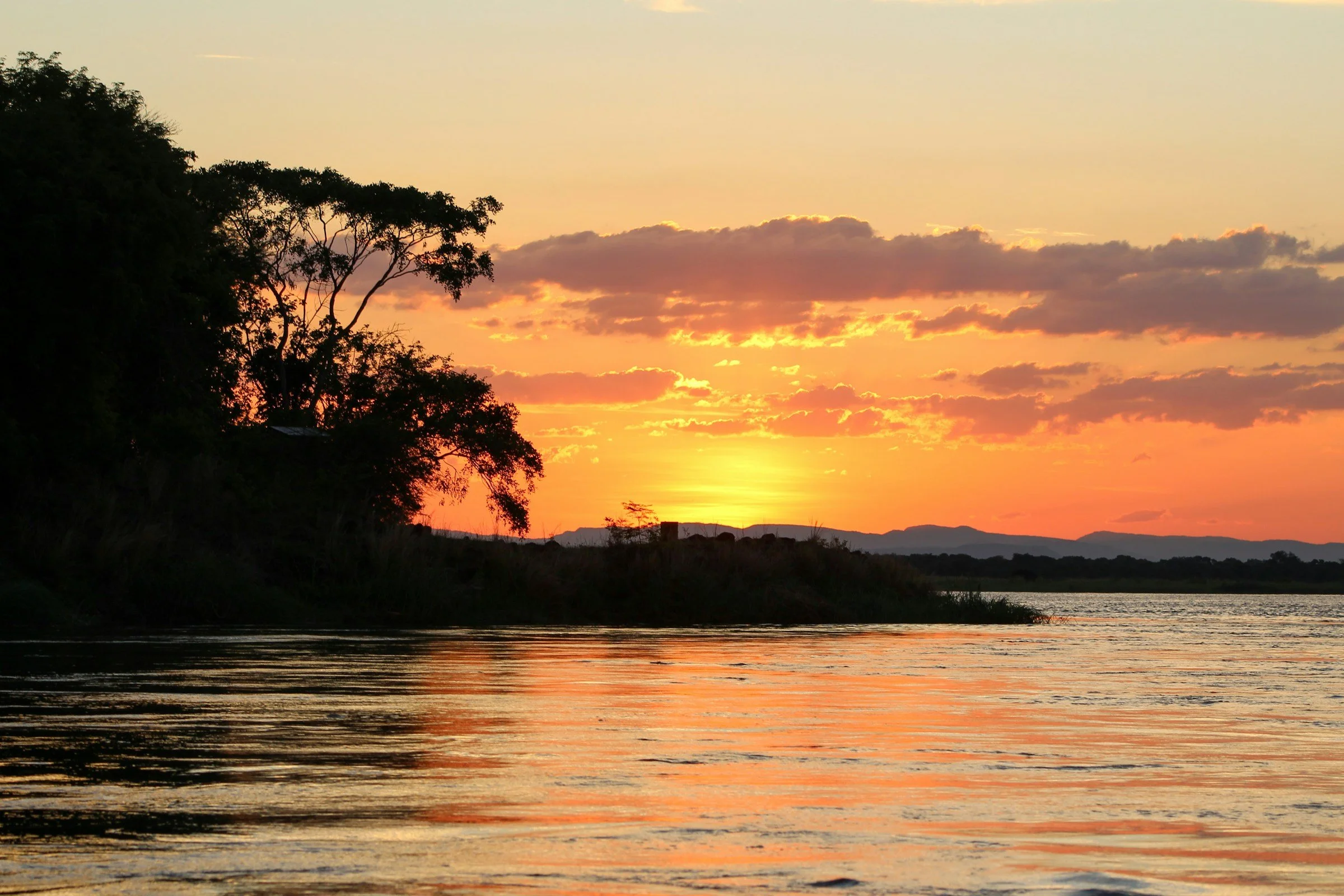 Sunset over the Zambezi river with silhouetted trees and mountains in the distance, orange and pink sky, calm water reflecting the colors.