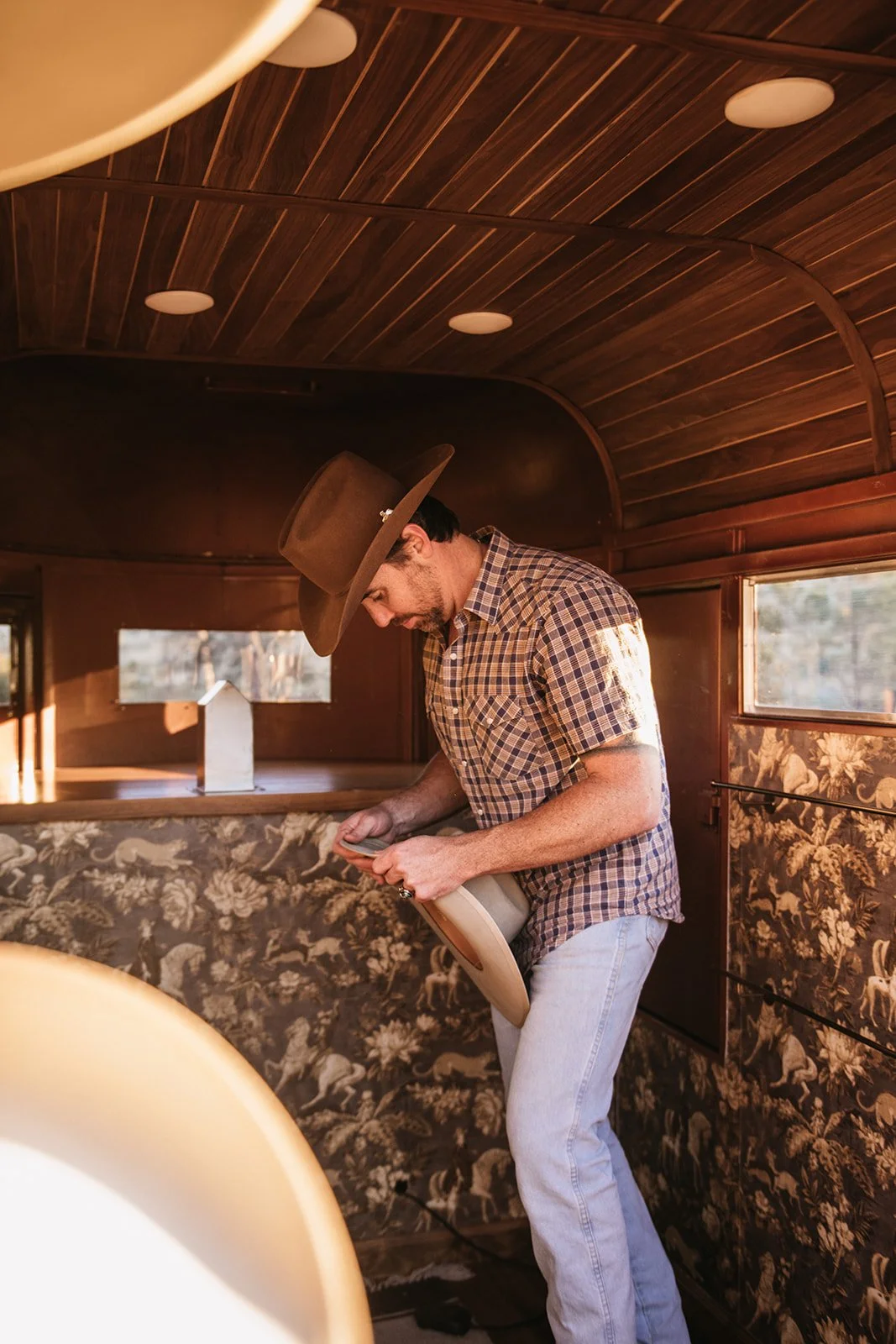 A man wearing a cowboy hat, plaid shirt, and light jeans, standing on a vintage bus with wood-paneled ceiling and floral wallpaper, looking at his phone.