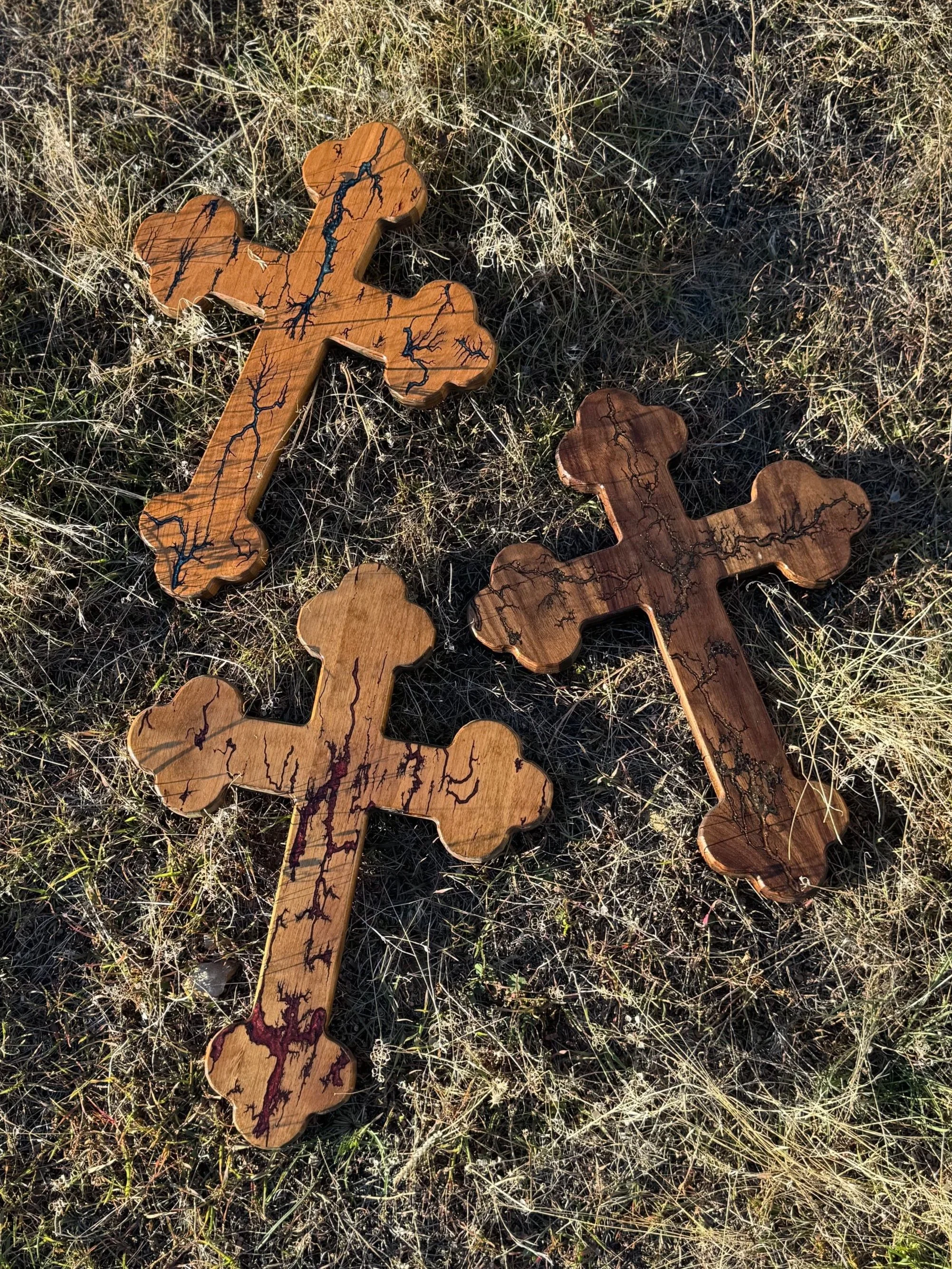 Three wooden crosses with black vein-like patterns lying on dry grass and small plants.