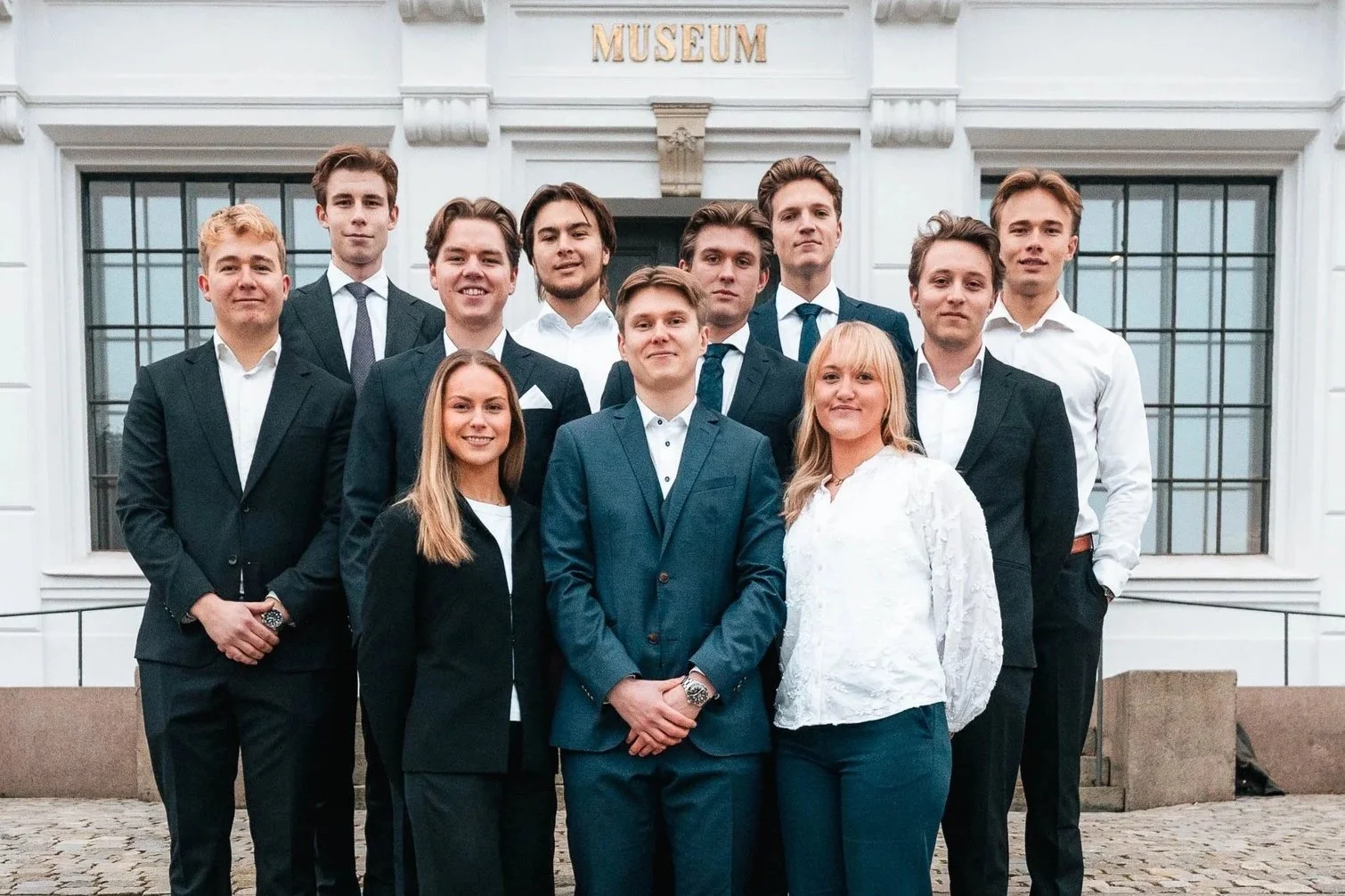 Bilde av Econos Invest: en gruppe på elleve unge studenter. De står på en trapp utenfor en stor, hvit bygning med store vinduer og en mørkegrønn dør, kledd i profesjonelle og formelle antrekk.