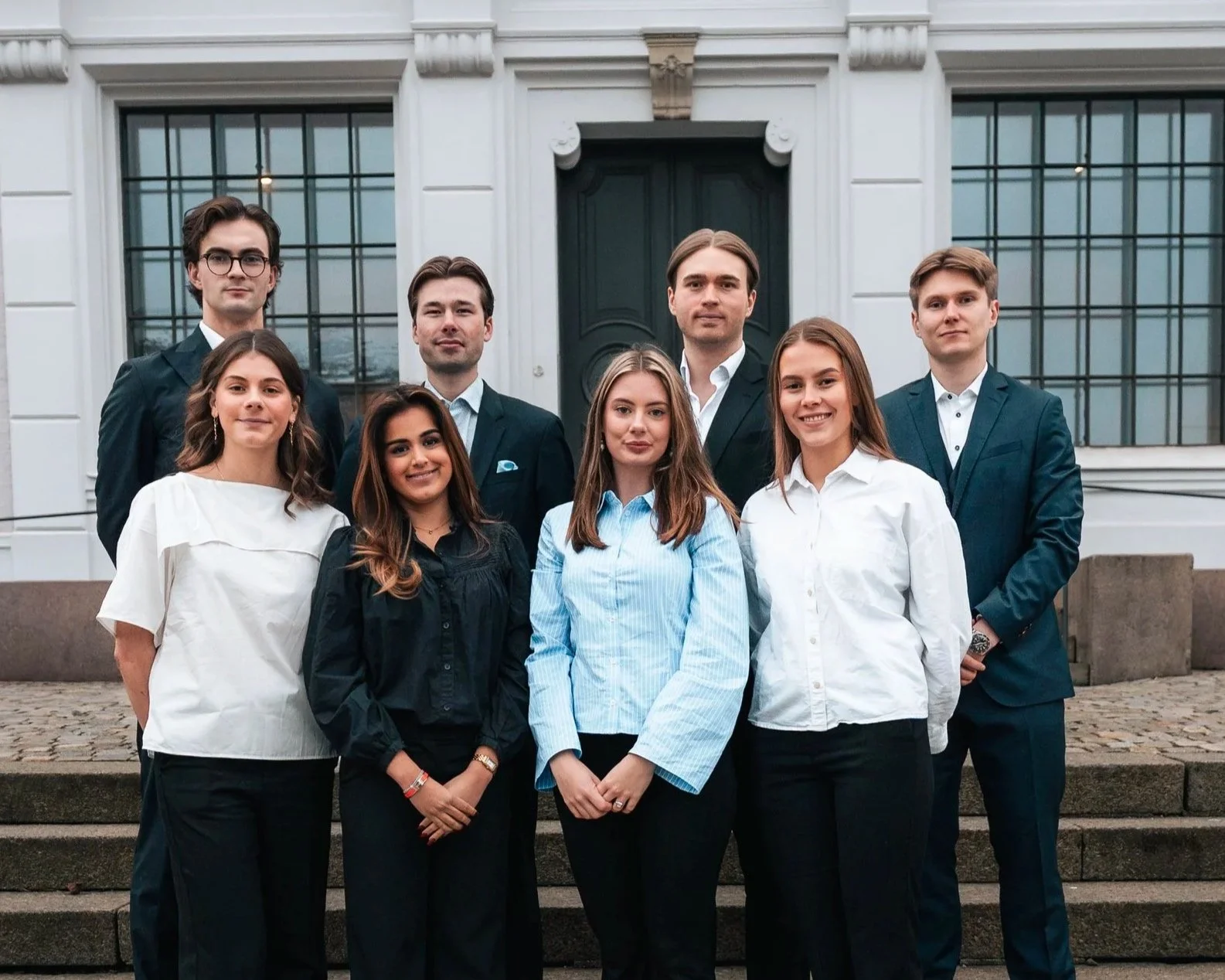 Bilde av styret i Econos: en gruppe på åtte unge studenter, fire menn og fire kvinner. De står på en trapp utenfor en stor, hvit bygning med store vinduer og en mørkegrønn dør, kledd i profesjonelle og formelle antrekk.