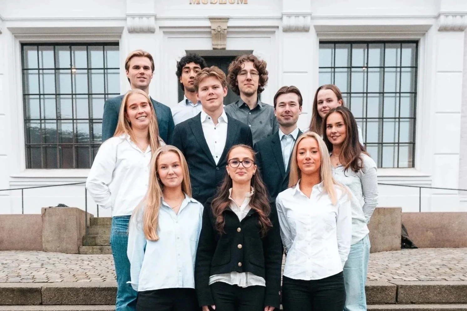 Bilde av karrieredagskomiteen i Econos: en gruppe på 11 unge studenter. De står på en trapp utenfor en stor, hvit bygning med store vinduer og en mørkegrønn dør, kledd i profesjonelle og formelle antrekk.